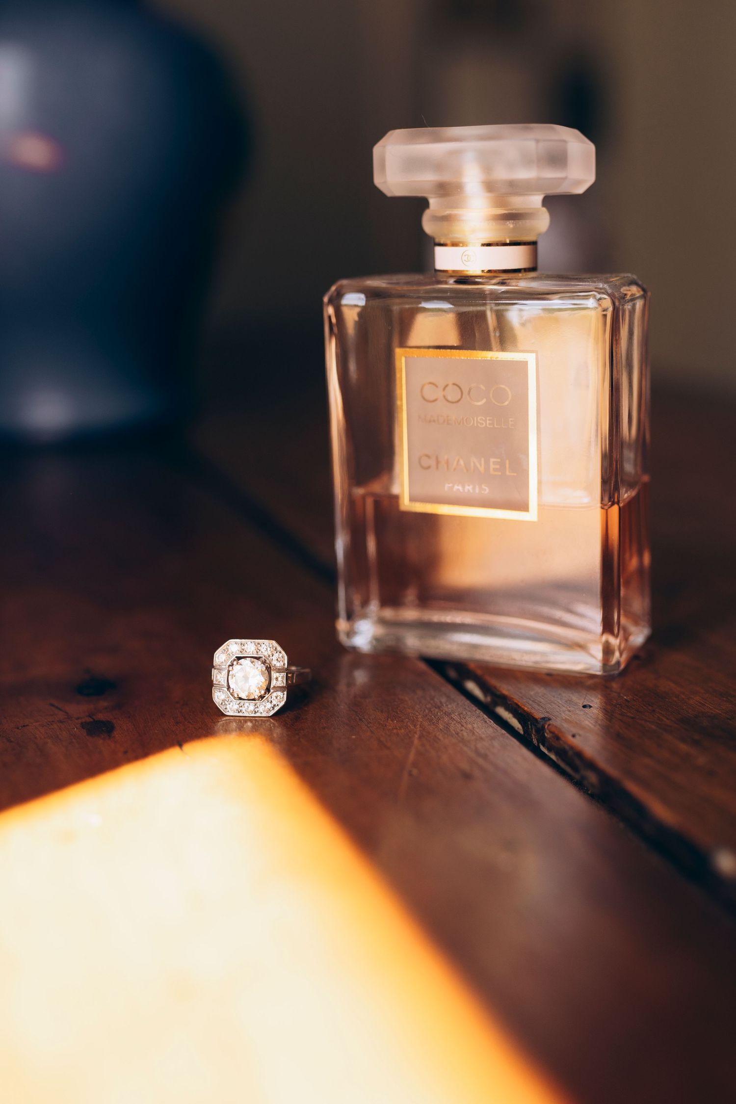 Bouteille de parfum Coco Mademoiselle Chanel à côté d'une bague halo en diamant sur une surface en bois dans une lumière chaude.