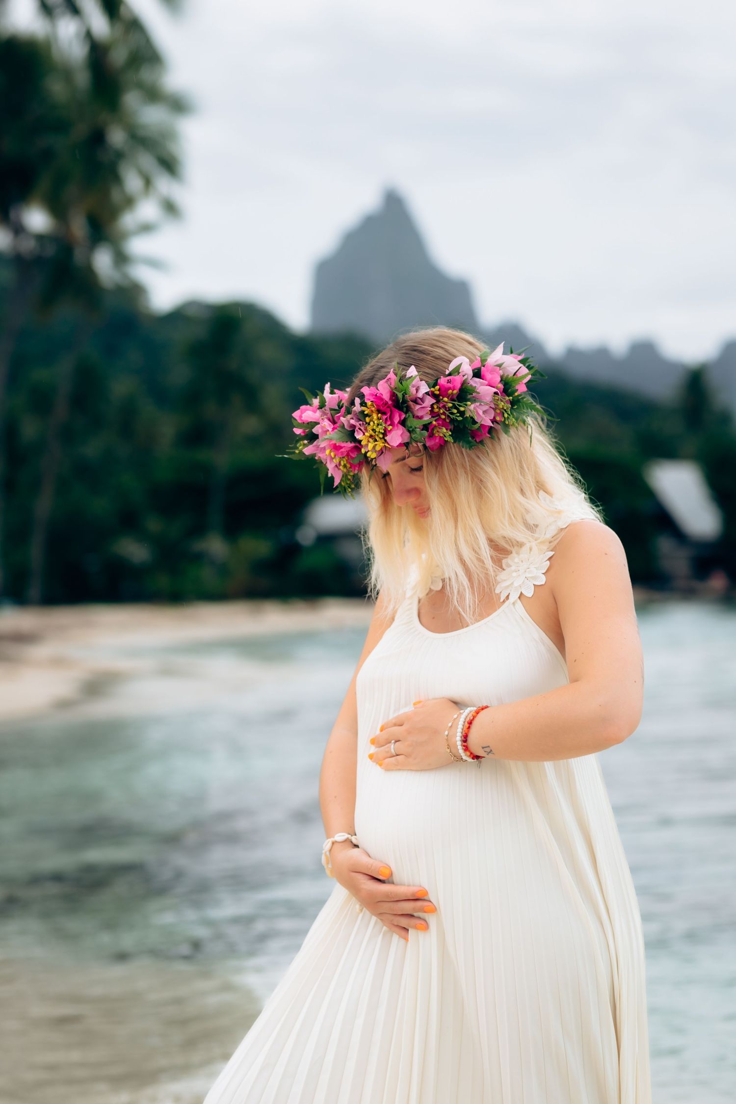 Femme enceinte en robe blanche et couronne florale rose berçant son ventre de bébé au bord d'une plage tropicale.