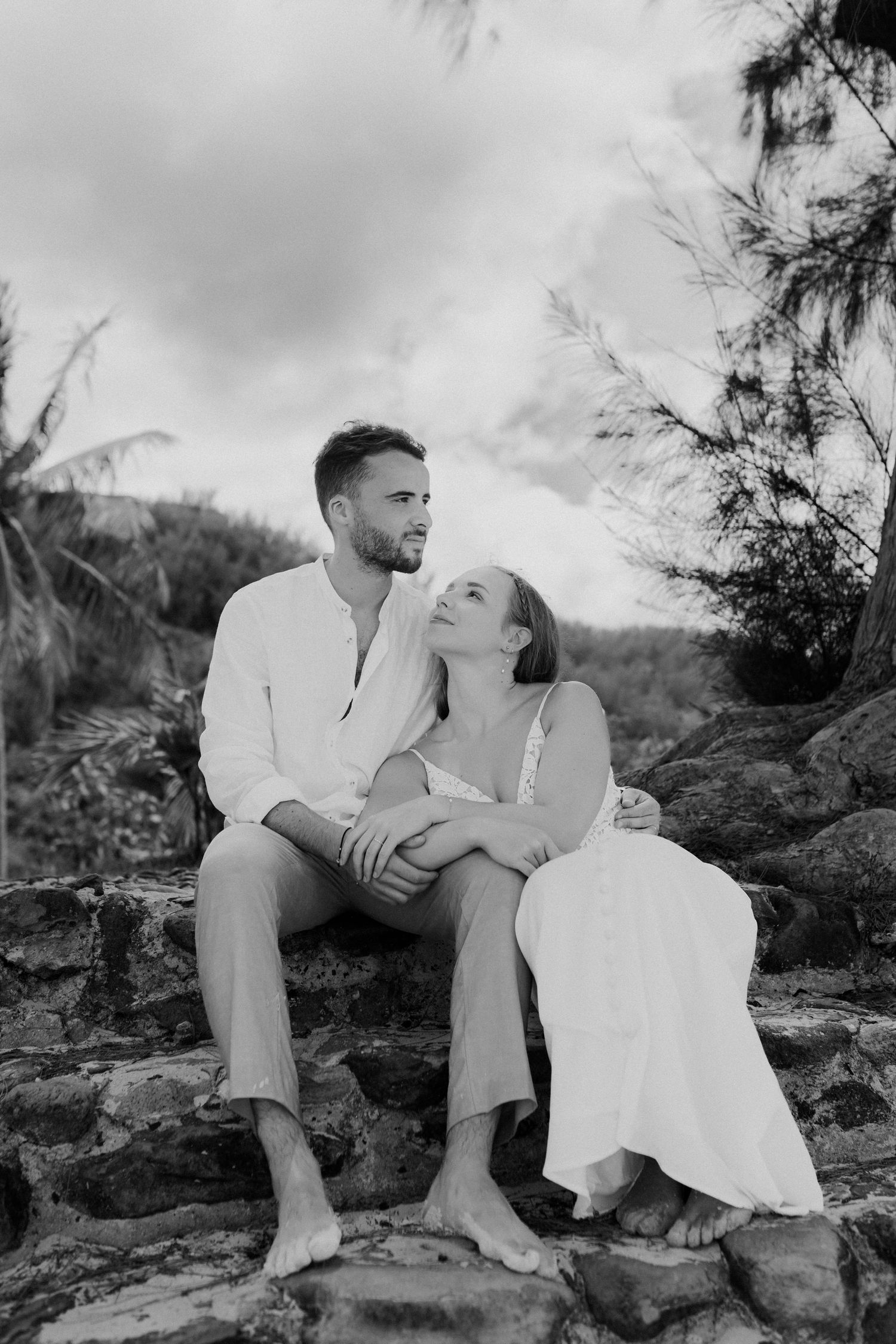 Black and white photo of couple sitting on rocks embracing in tropical outdoor setting.