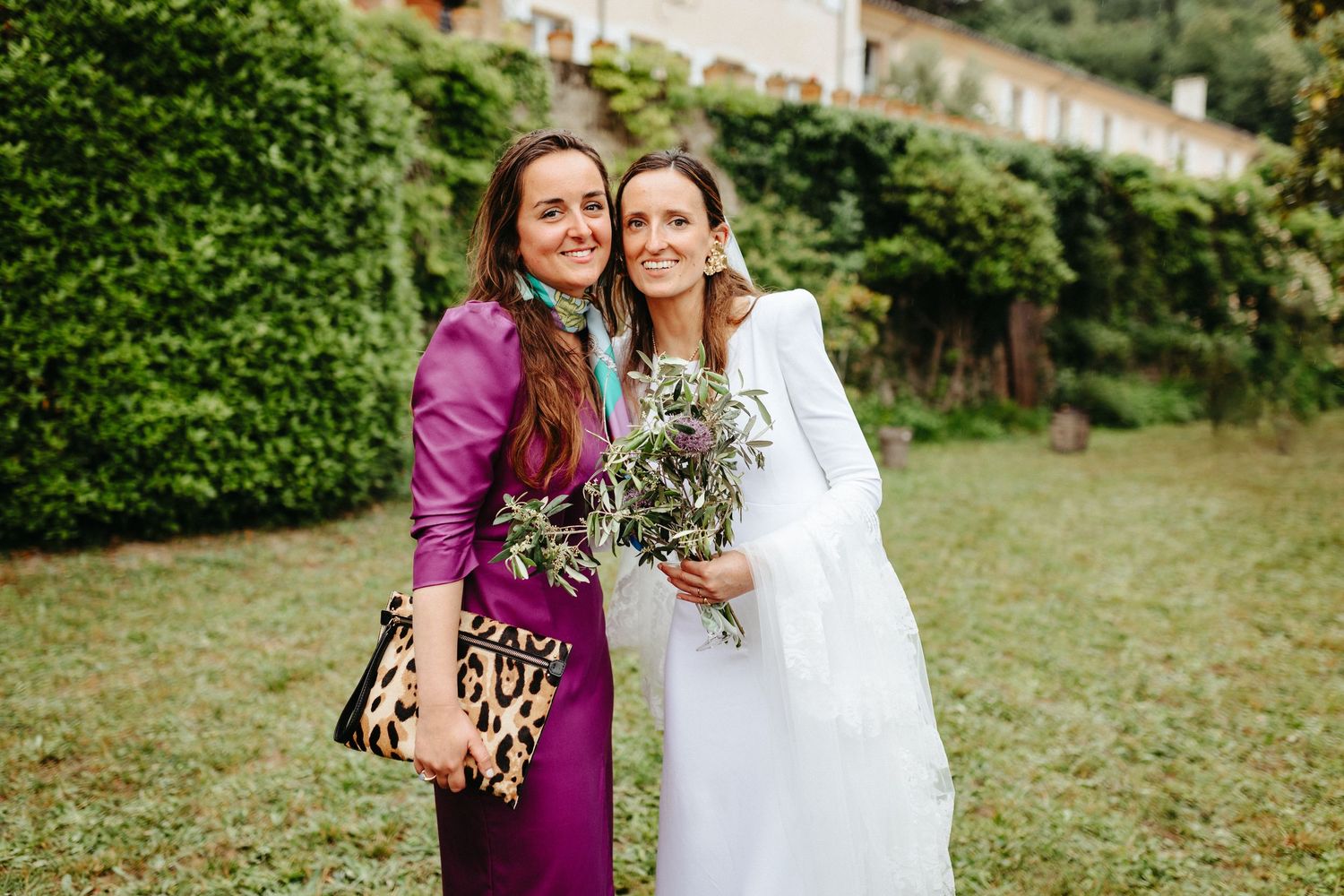 Deux soeurs souriantes, l'une en robe de mariée blanche tenant un bouquet de verdure, l'autre en robe violette avec un sac léopard