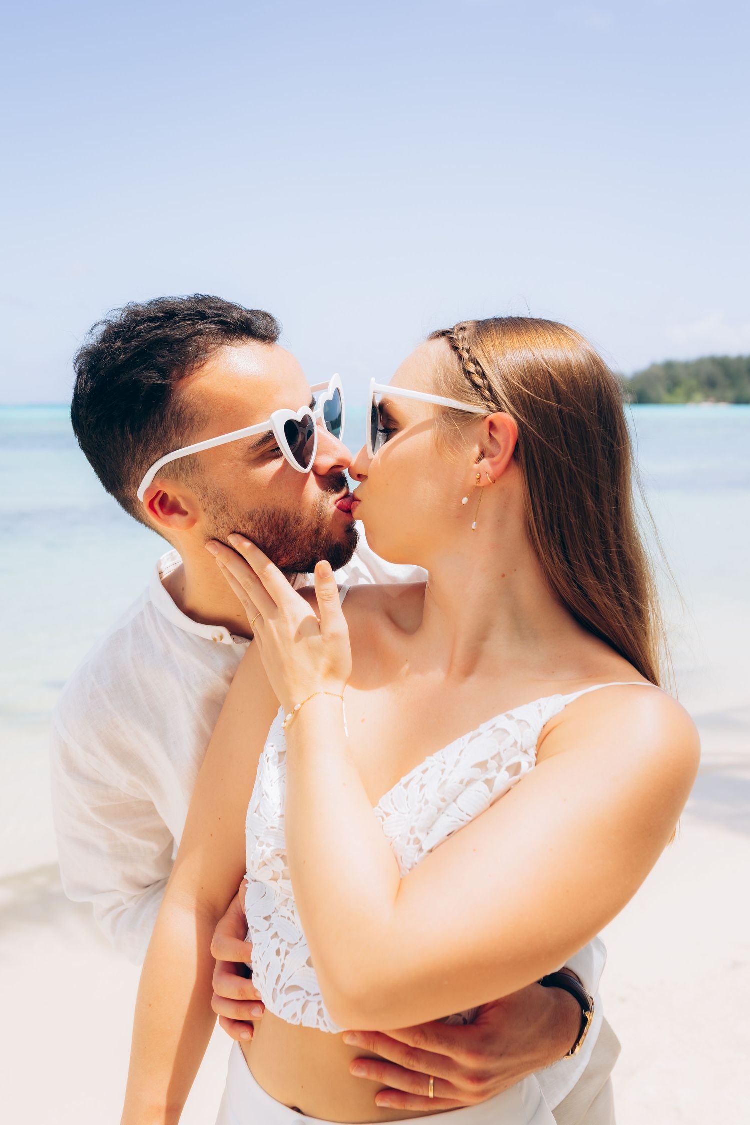 Couple en tenue blanche s'embrassant sur une plage tropicale portant des lunettes de soleil en forme de cœur.