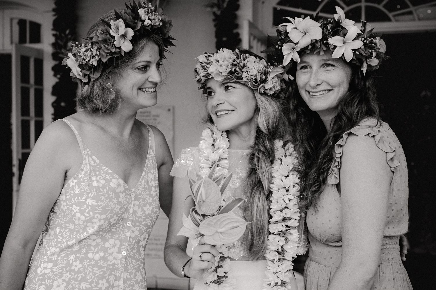 Trois femmes portant des couronnes florales sourient ensemble lors d'une célébration de mariage tropical en noir et blanc.