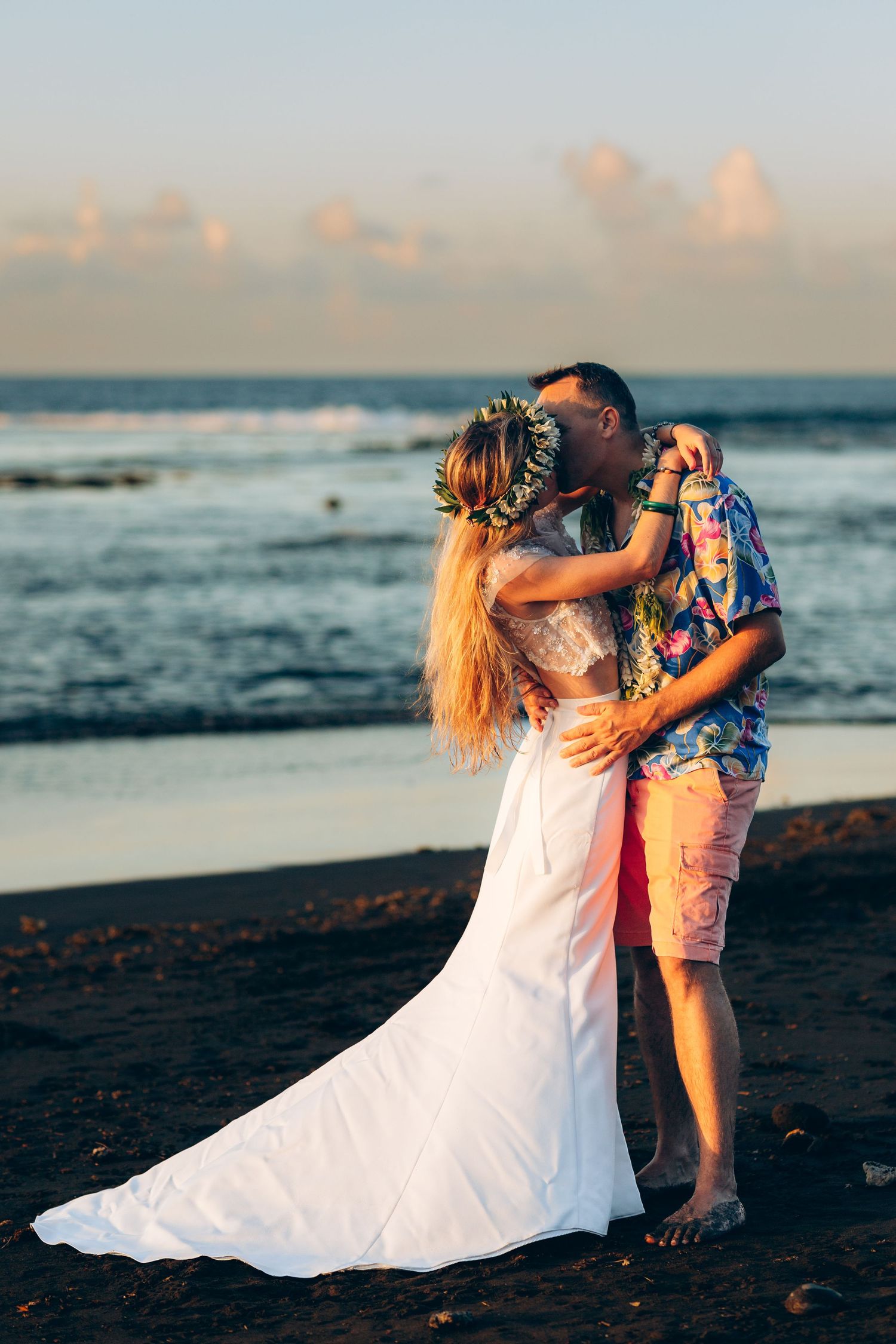 Couple partageant un baiser sur une plage de sable noir au coucher du soleil, mariée en robe blanche et couronne florale.