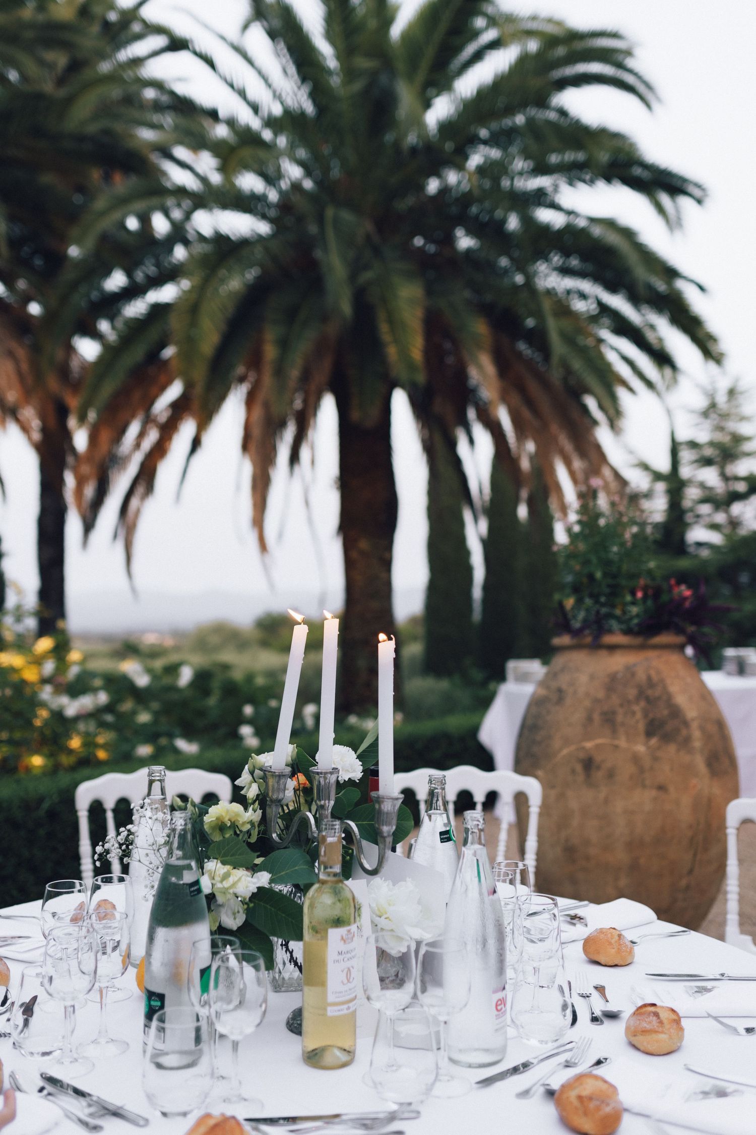 Cadre de table de mariage en plein air élégant avec des bougies, du vin et des fleurs, encadré par des palmiers luxuriants.