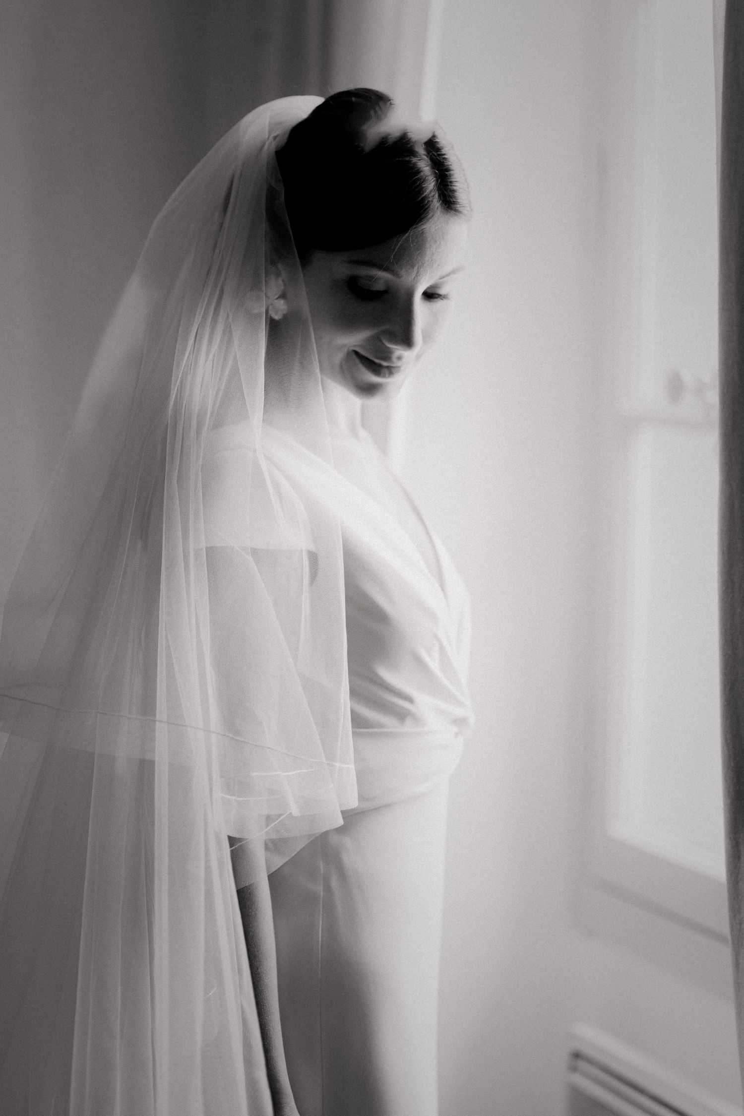 Elegant bride in white gown and veil smiling by window in black and white photo.