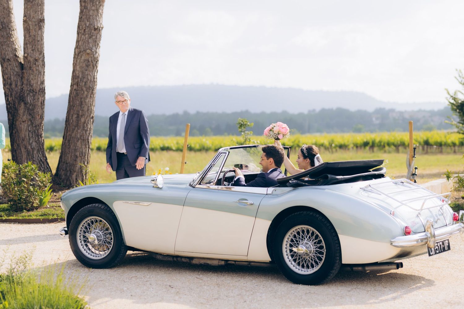 Couple roulant dans une voiture de sport décapotable argentée vintage dans un lieu de mariage viticole avec des montagnes pittoresques.