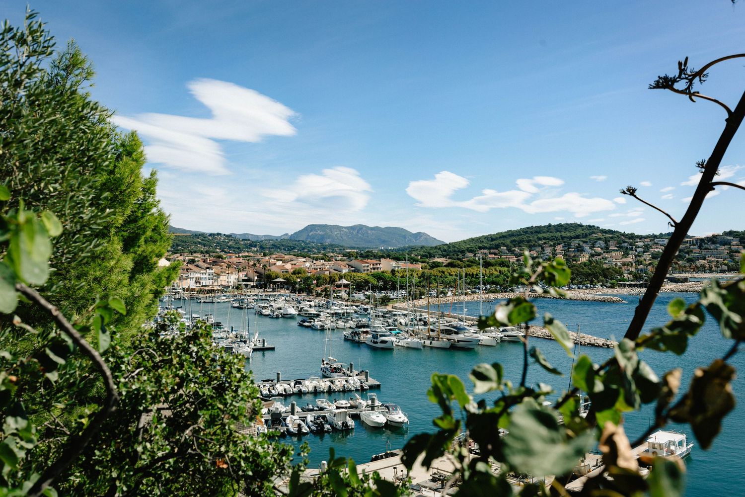 Port méditerranéen pittoresque avec des voiliers amarrés, une verdure luxuriante et une toile de fond de montagne sous un ciel bleu.