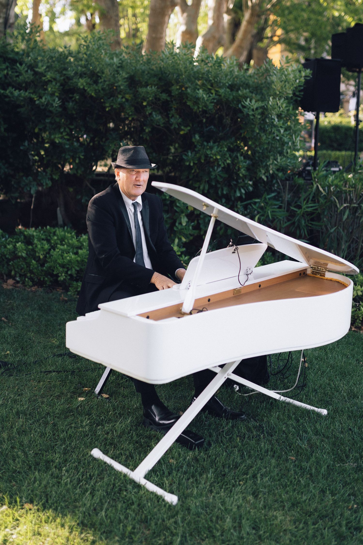 Homme en costume noir et fedora jouant d'un piano à queue blanc à l'extérieur sur de l'herbe verte avec des haies luxuriantes derrière lui.