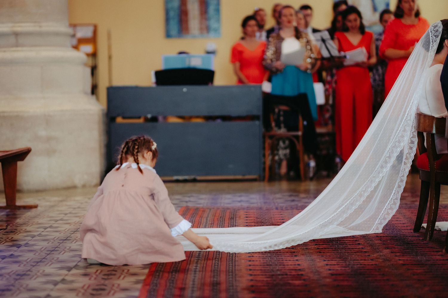 Une fille d'honneur agenouillée sur un tapis rouge en arrangeant le long voile d'une mariée à l'intérieur d'une église toulonnaise