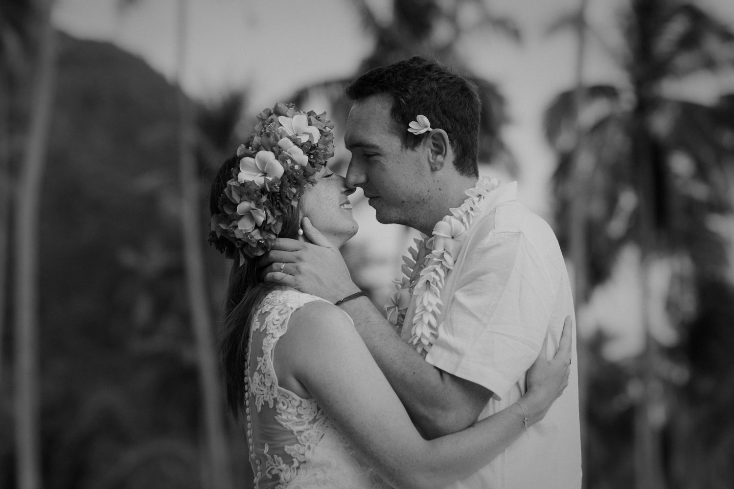 Gros plan noir et blanc de la mariée et du marié partageant un baiser romantique avec des leis floraux.