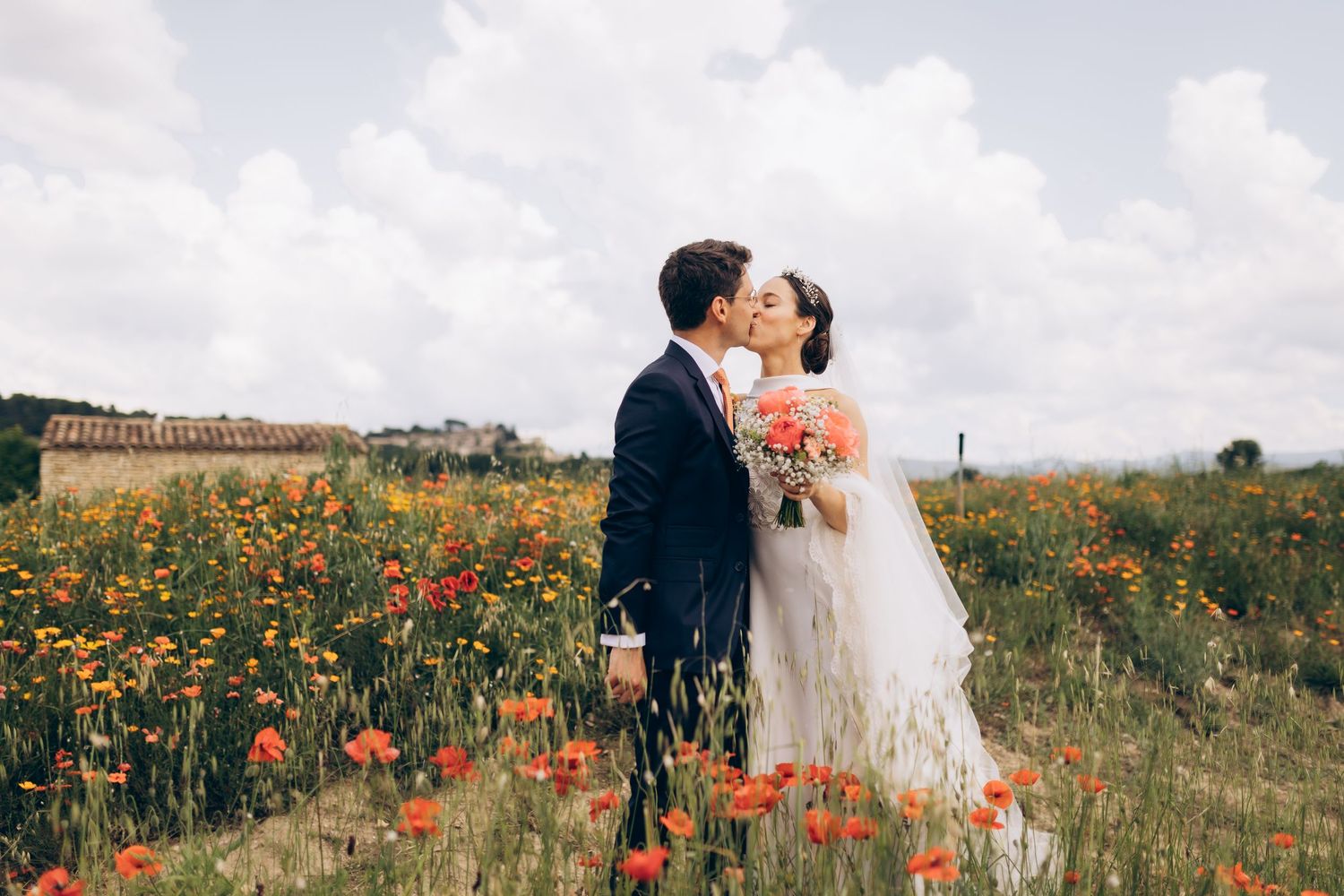 Couple s'embrassant dans un champ de coquelicots vibrant, mariée en robe blanche tenant un bouquet, marié en costume bleu marine