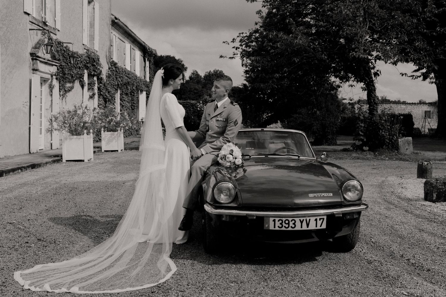 Photo de mariage en noir et blanc de la mariée et du marié posant avec une voiture décapotable vintage à l'extérieur d'un château français.