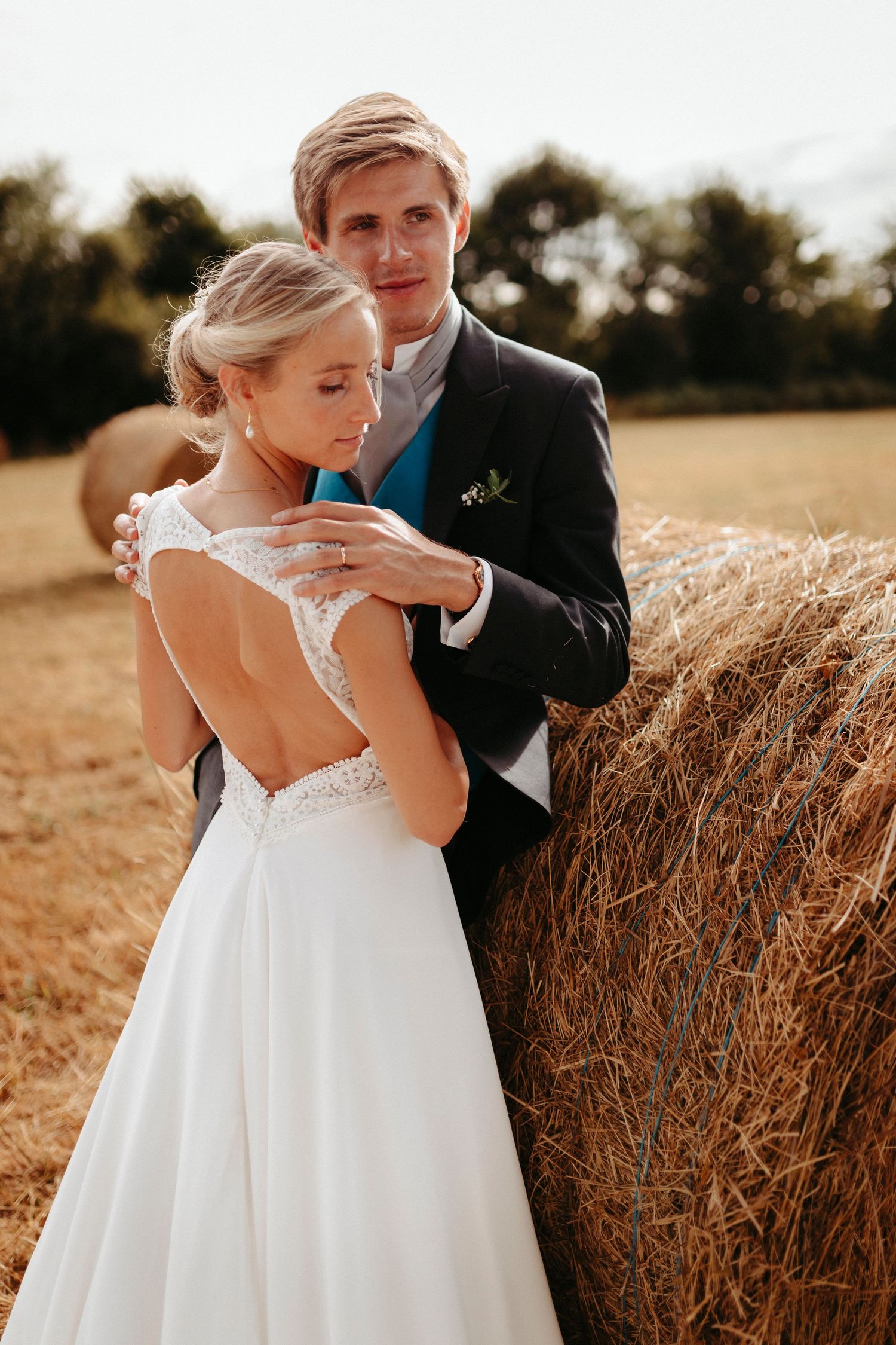 La mariée en robe blanche dos nu et le marié en costume bleu marine s'embrassent près d'une balle de foin dans un champ doré ensoleillé.
