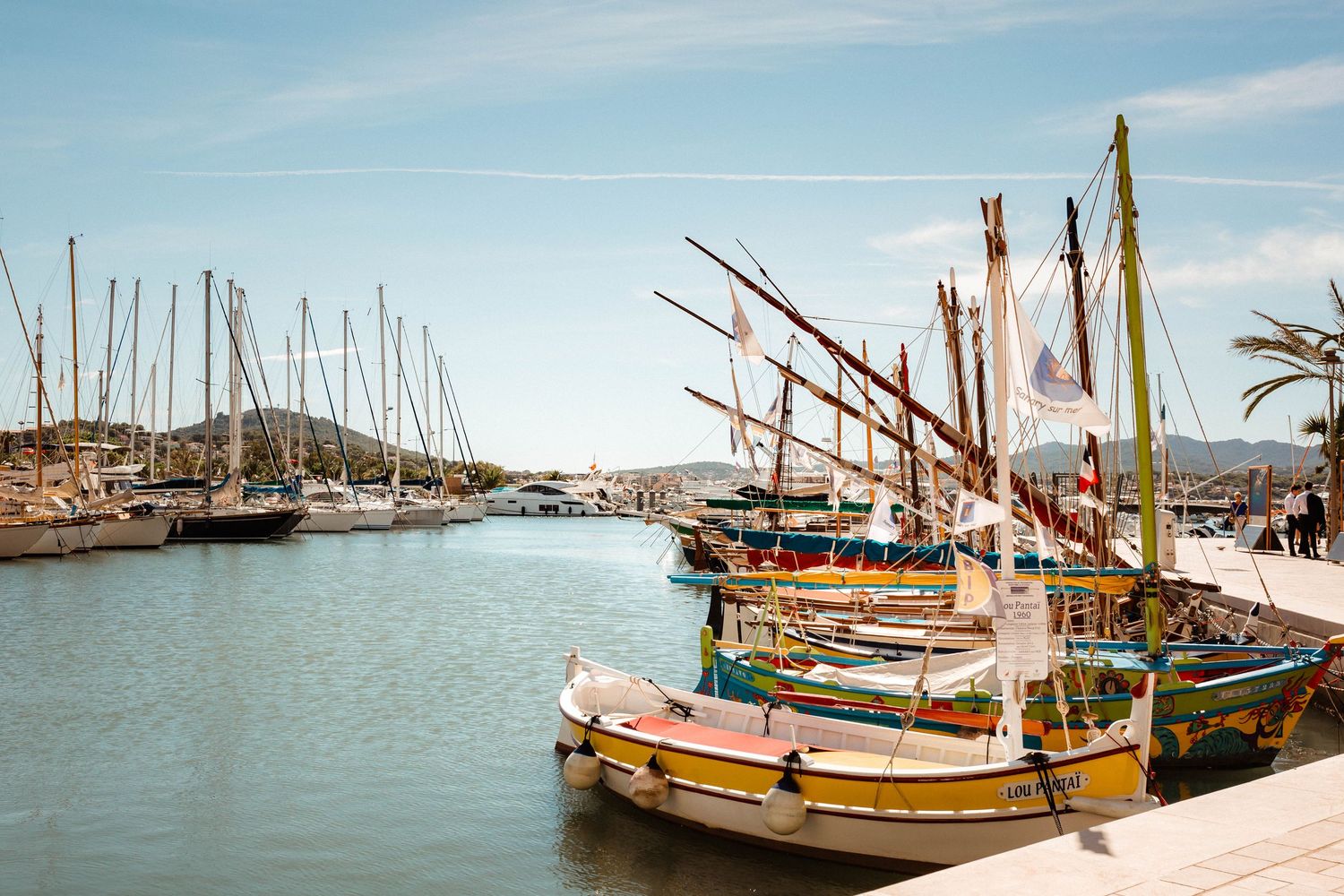 Des bateaux traditionnels colorés amarrés dans un port méditerranéen ensoleillé avec des voiliers et des eaux bleues calmes en arrière-plan.