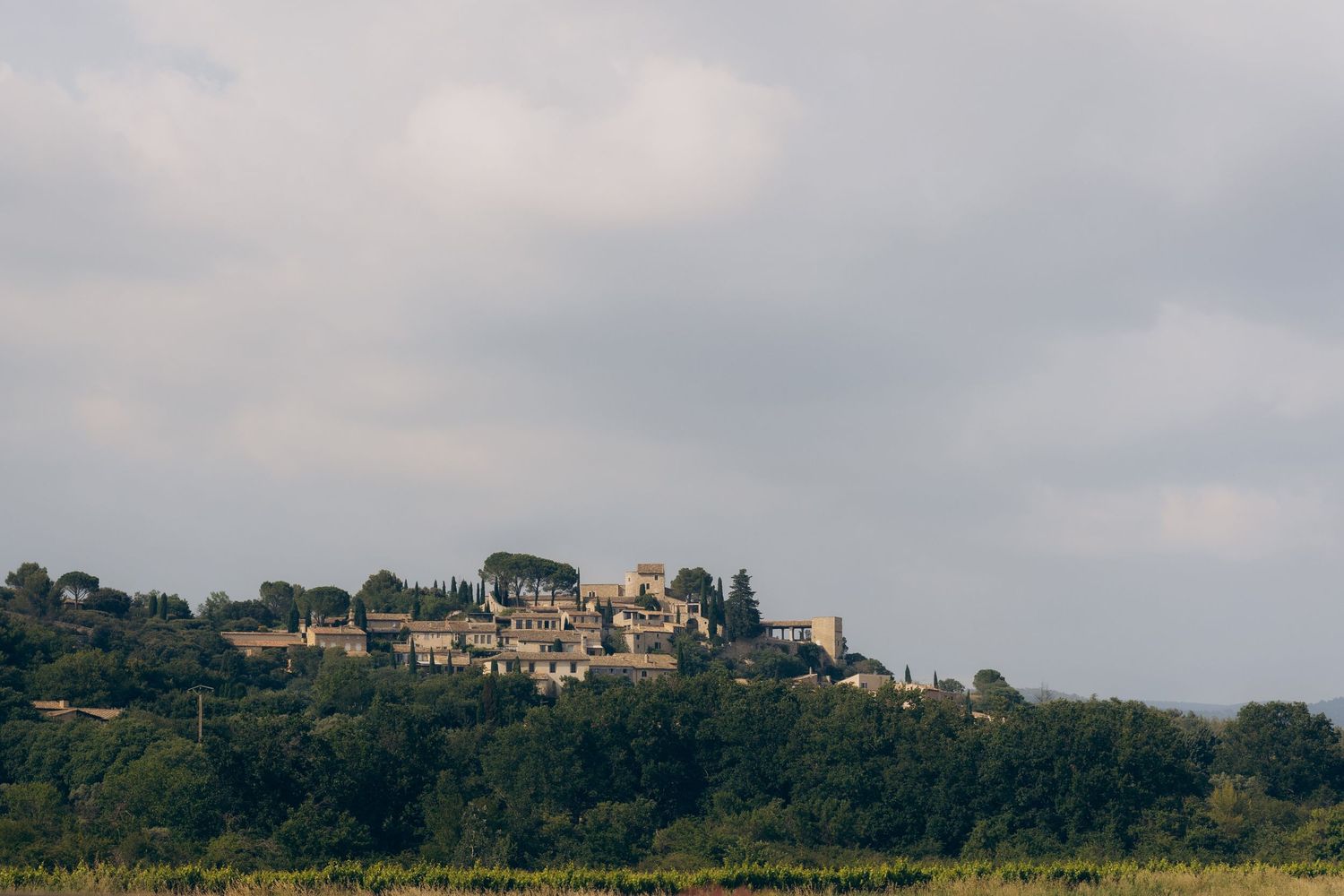 Un village provençal au sommet d'une colline entouré d'arbres verts luxuriants sous un ciel nuageux dans la campagne française.