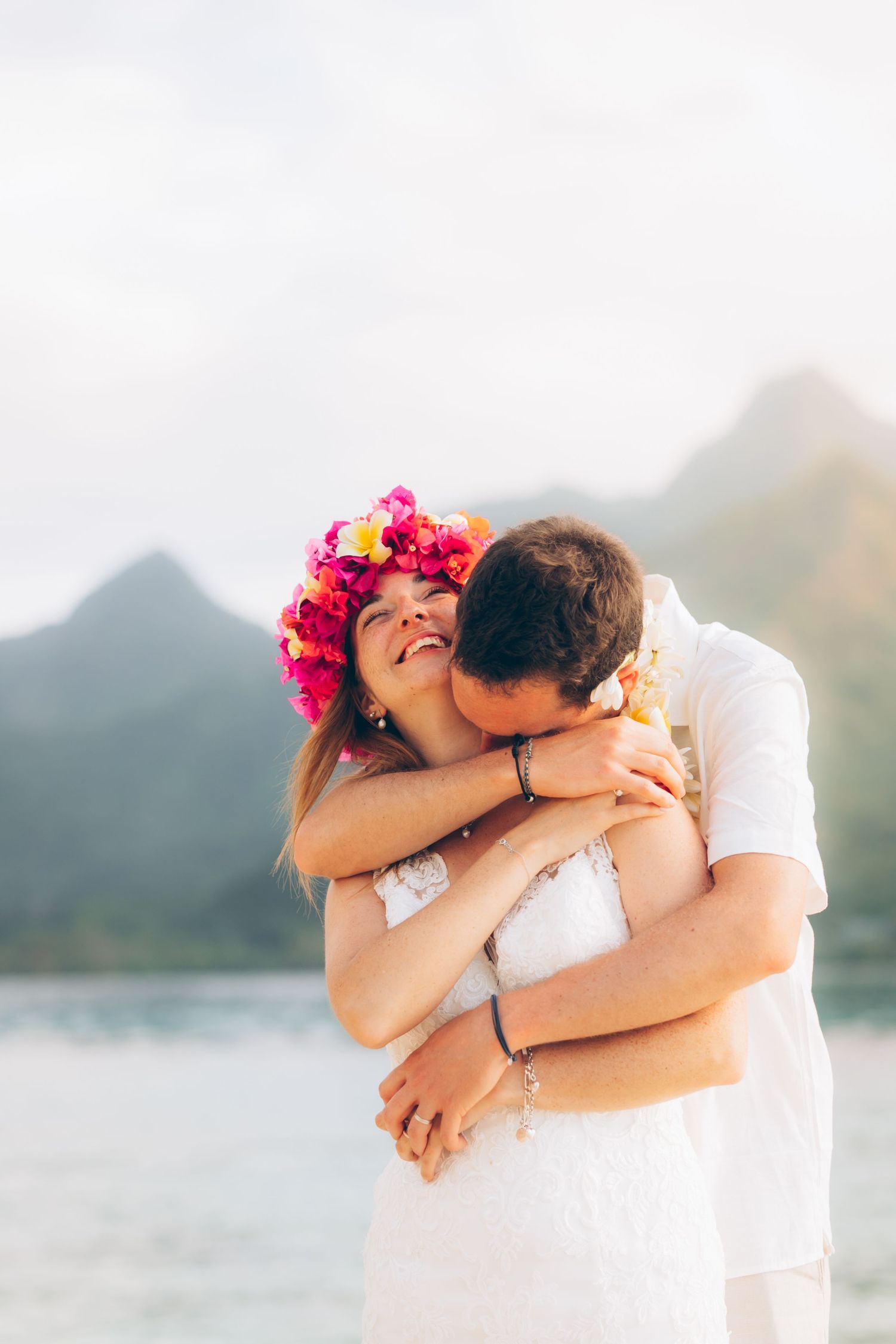 Une mariée souriante portant une couronne florale vibrante embrasse le marié avec une toile de fond de montagne.