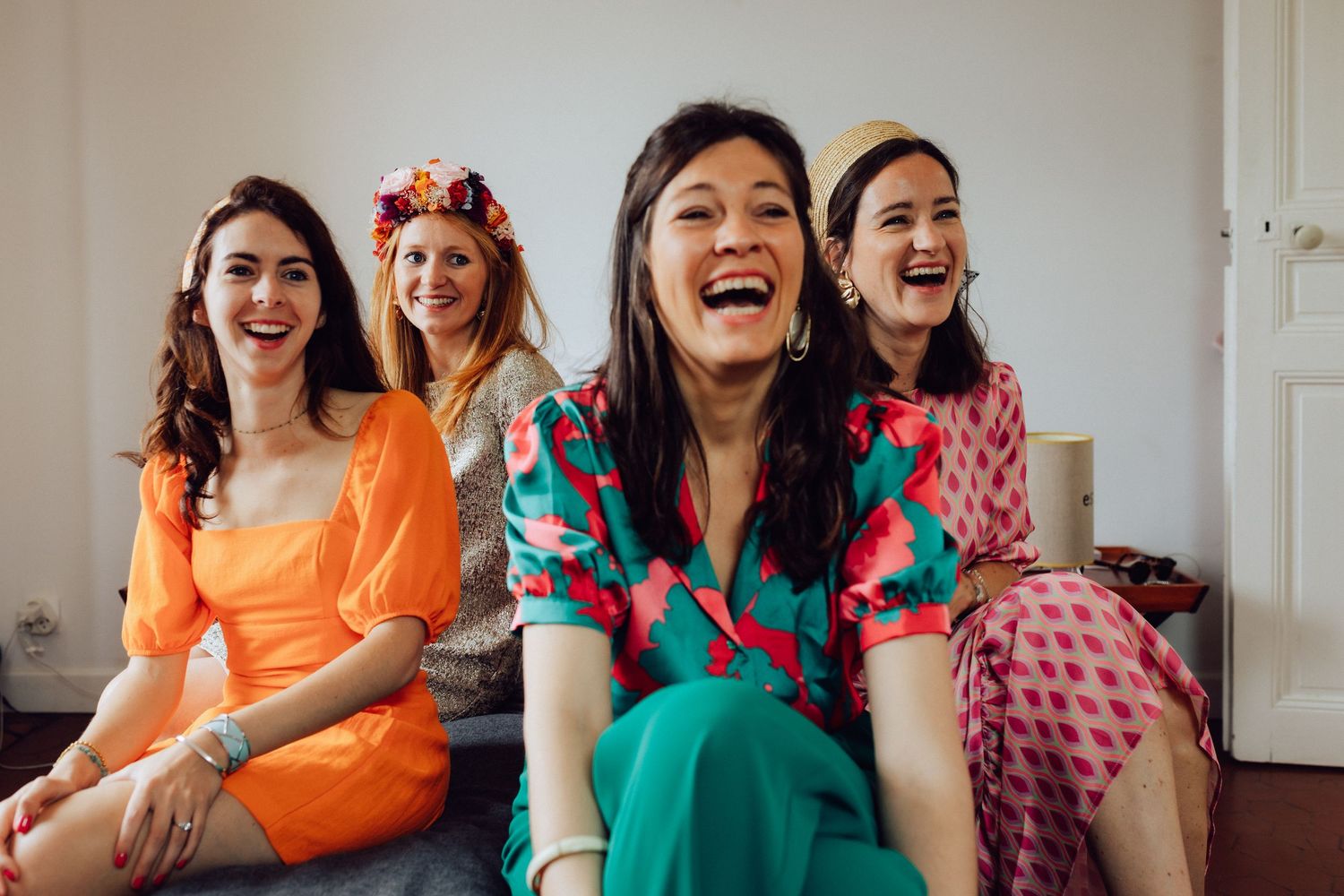 Quatre femmes rient et sont assises ensemble, portant des robes colorées aux motifs floraux orange, sarcelle et rose.