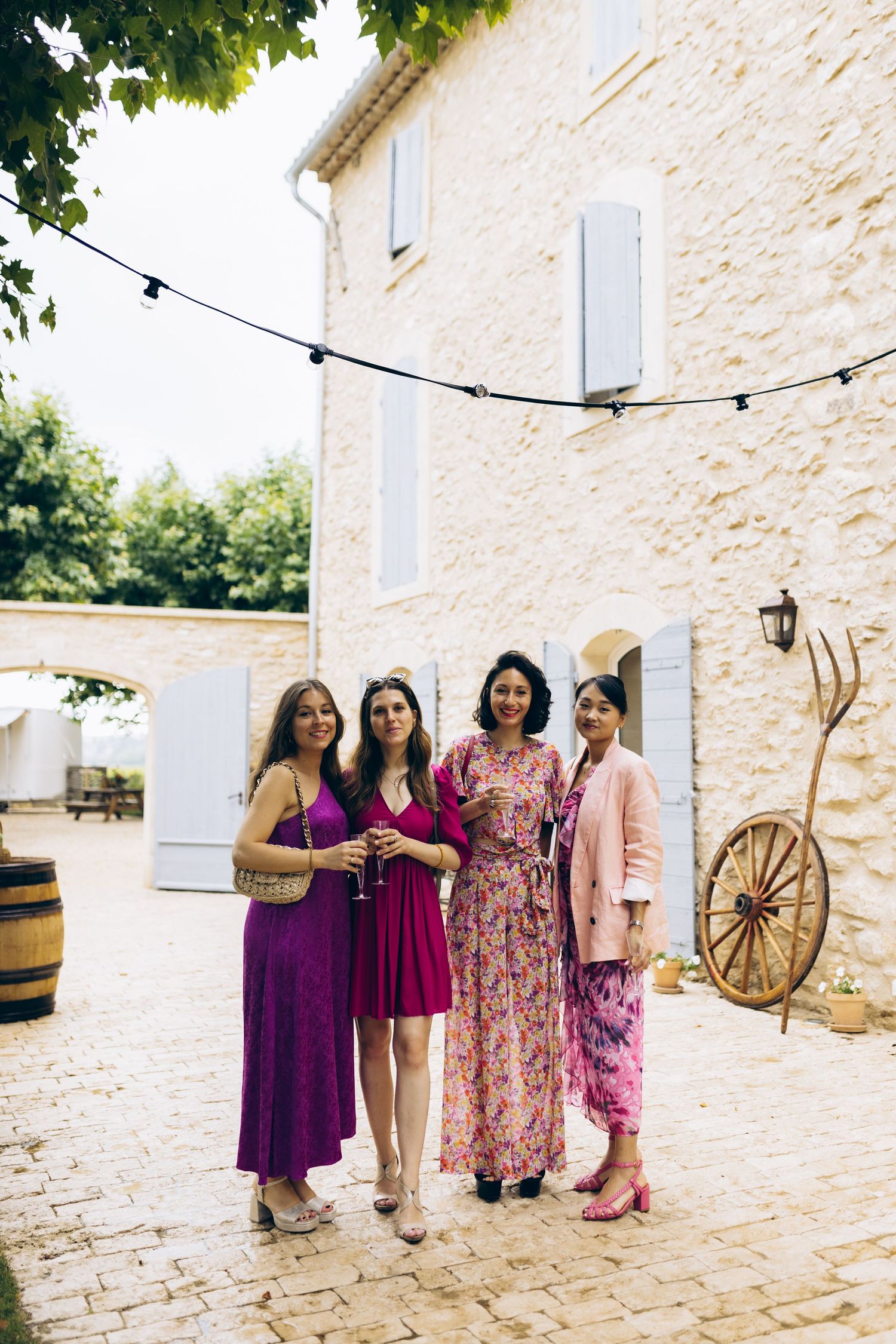 Quatre femmes en robes colorées posent ensemble dans un lieu de mariage en plein air avec des guirlandes lumineuses et un bâtiment en pierre