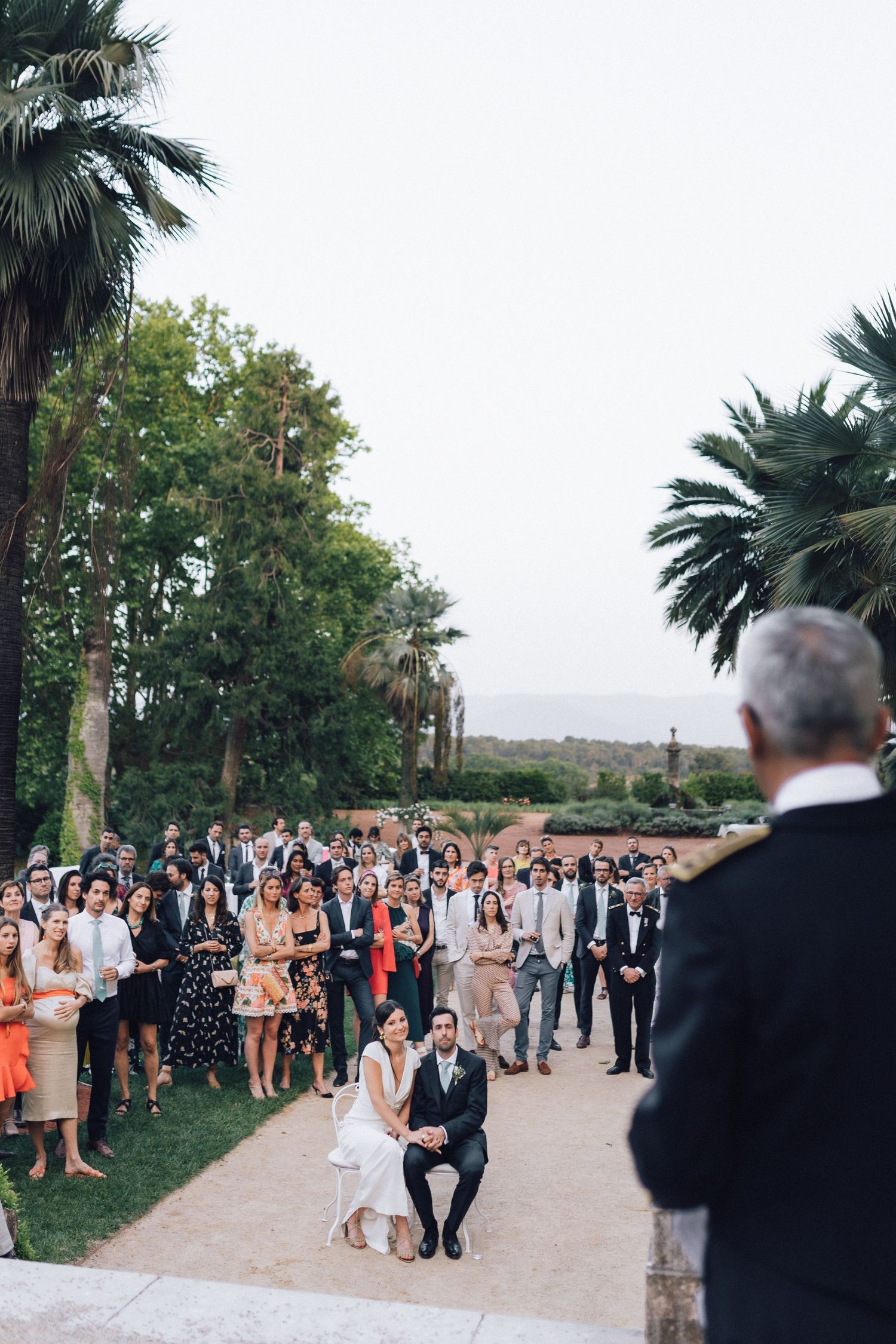 Cérémonie de mariage en plein air avec un couple assis devant les invités, entouré de palmiers et de verdure luxuriante.
