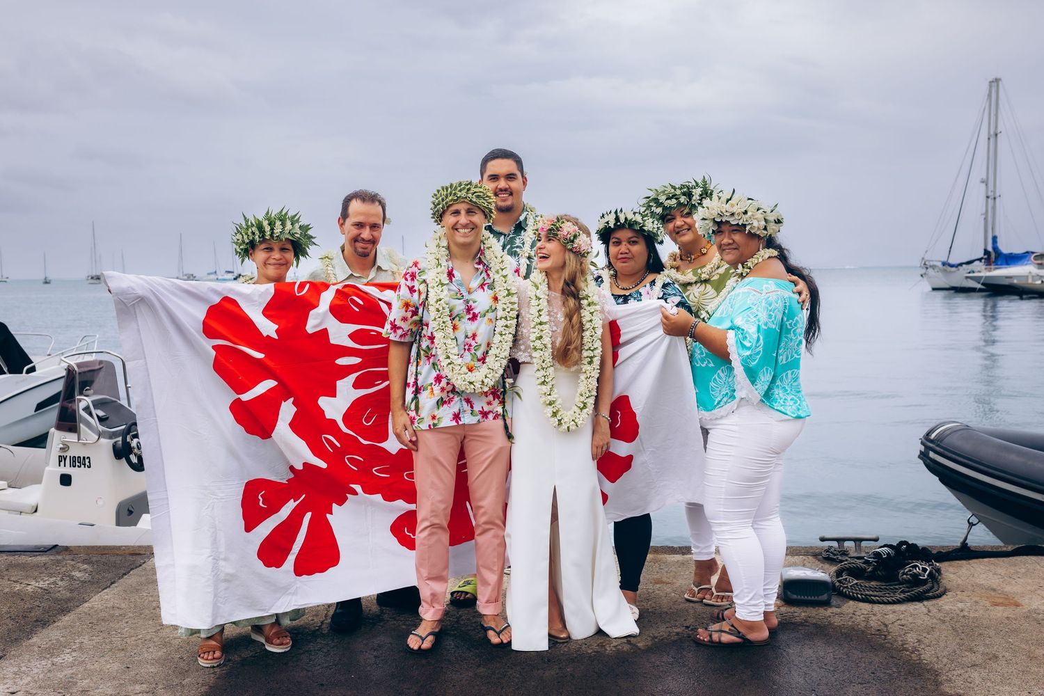 Les invités de mariage en tenue florale se mêlent près d'une marina avec des voiliers lors d'une célébration tropicale.