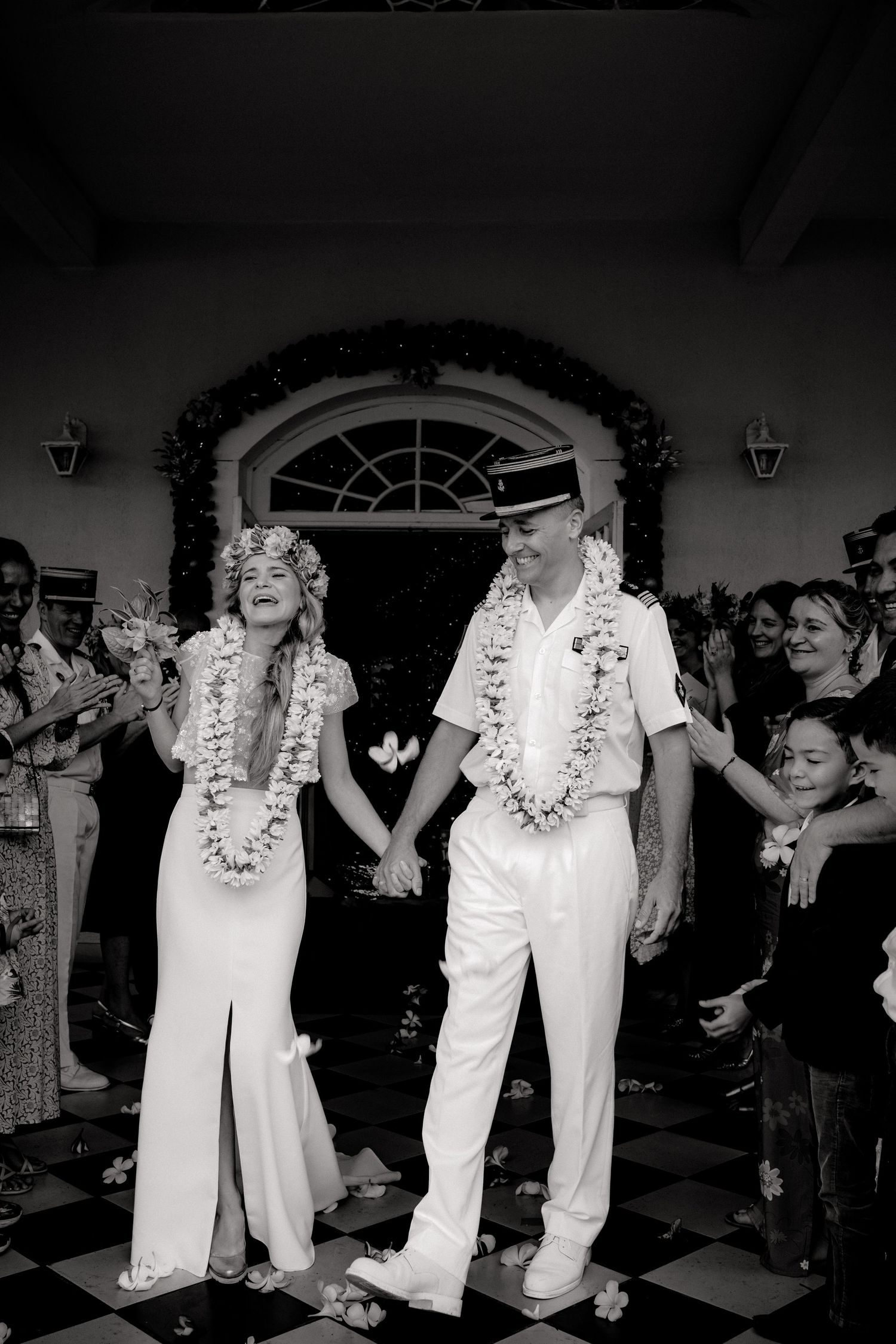 Photo en noir et blanc de jeunes mariés se tenant la main portant des leis alors que les invités célèbrent leur sortie de mariage.