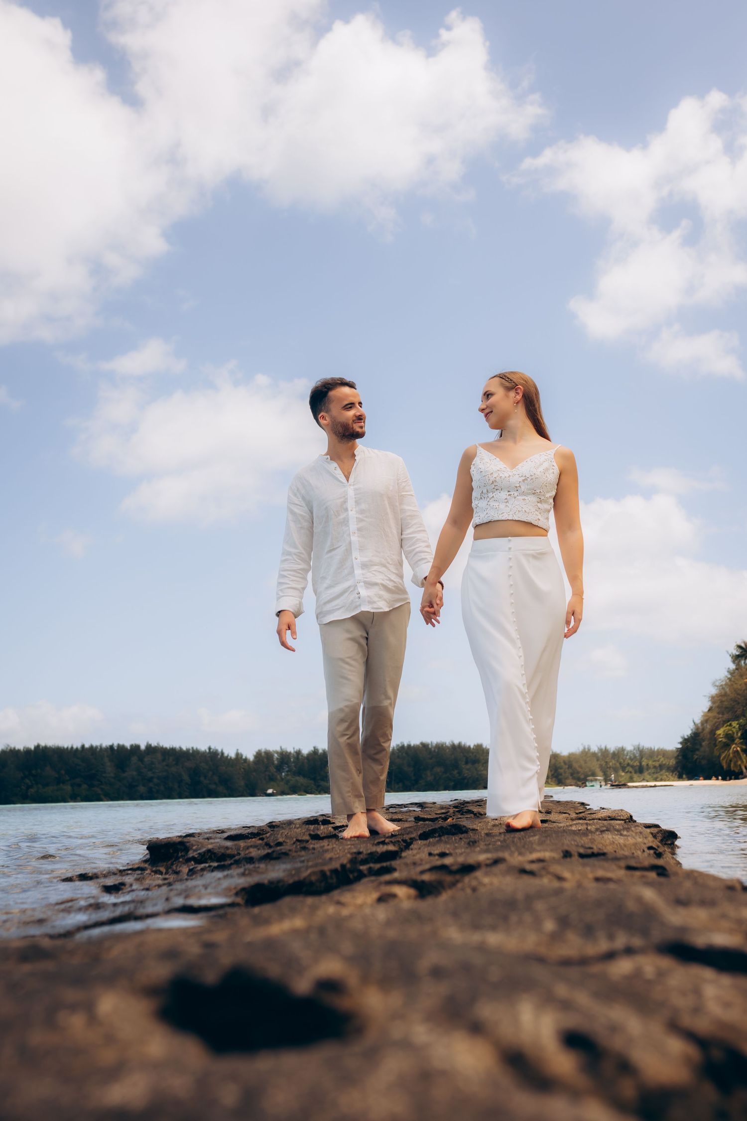 Couple en blanc se tenant la main marchant sur un rivage rocheux avec un ciel bleu et une vue sur l'océan.