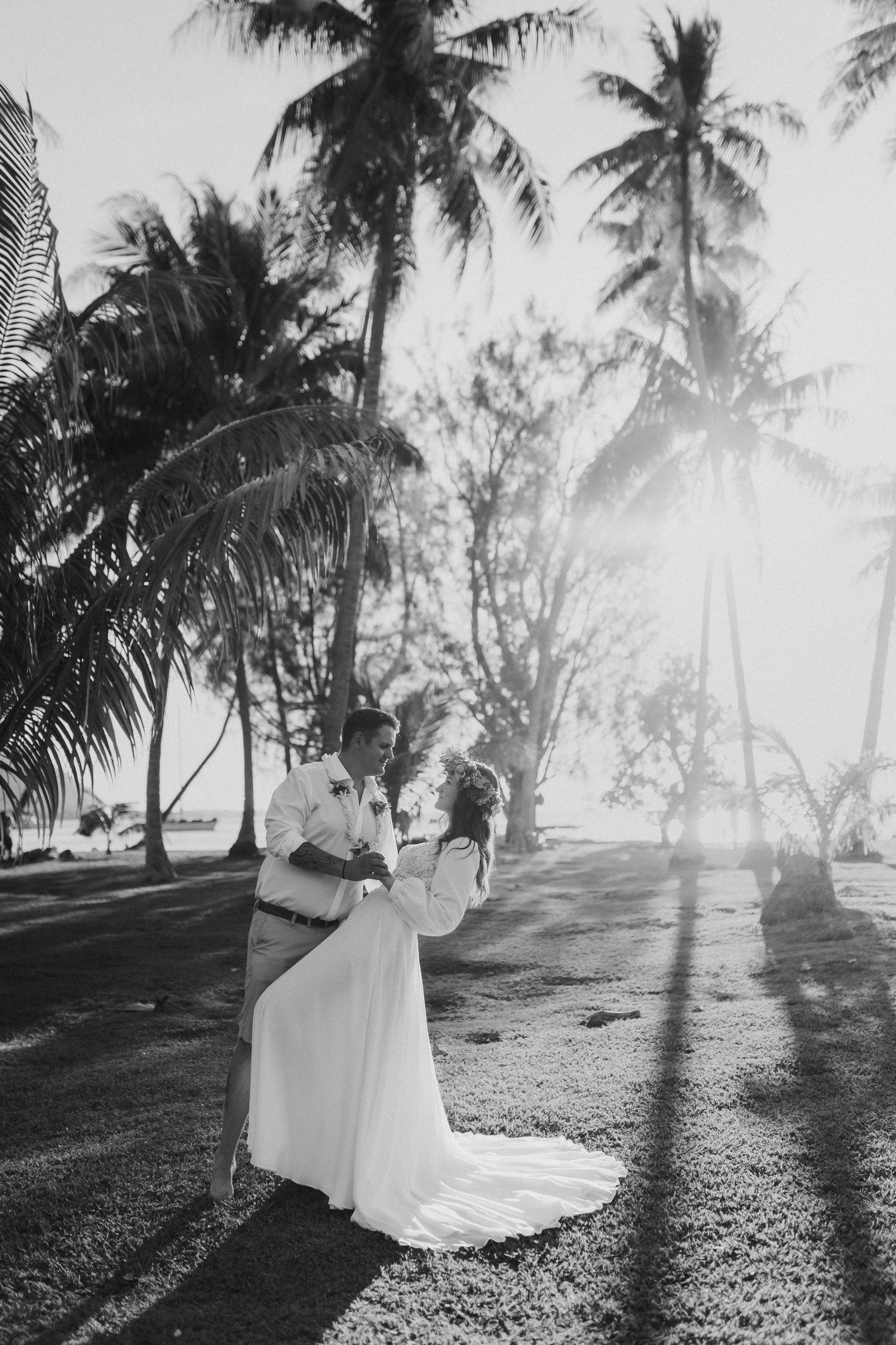 Photo en noir et blanc du marié soulevant la mariée parmi les palmiers avec la lumière du soleil.