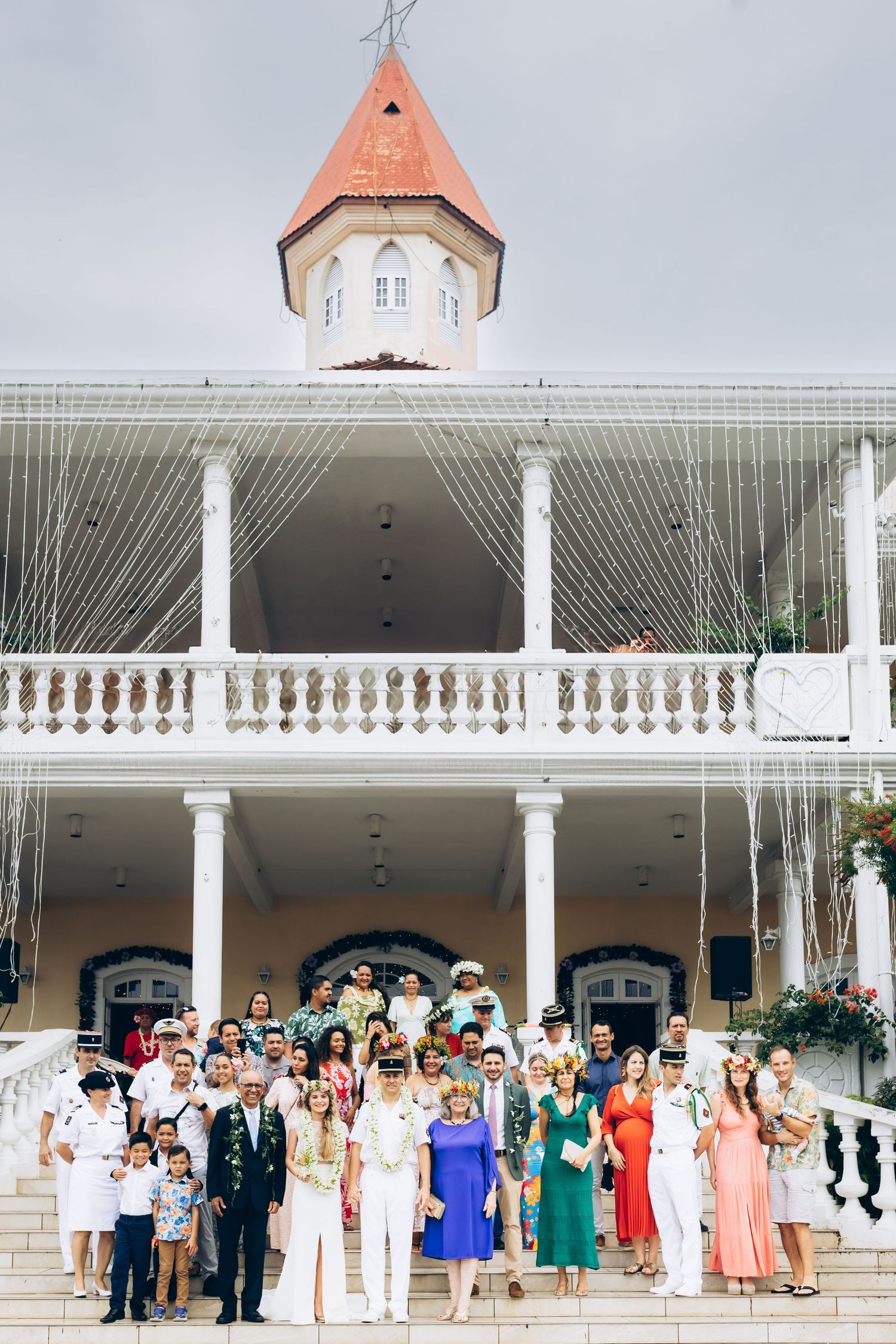 Fête de mariage et invités posent pour une photo de groupe sur les marches d'un élégant bâtiment colonial blanc avec tour.