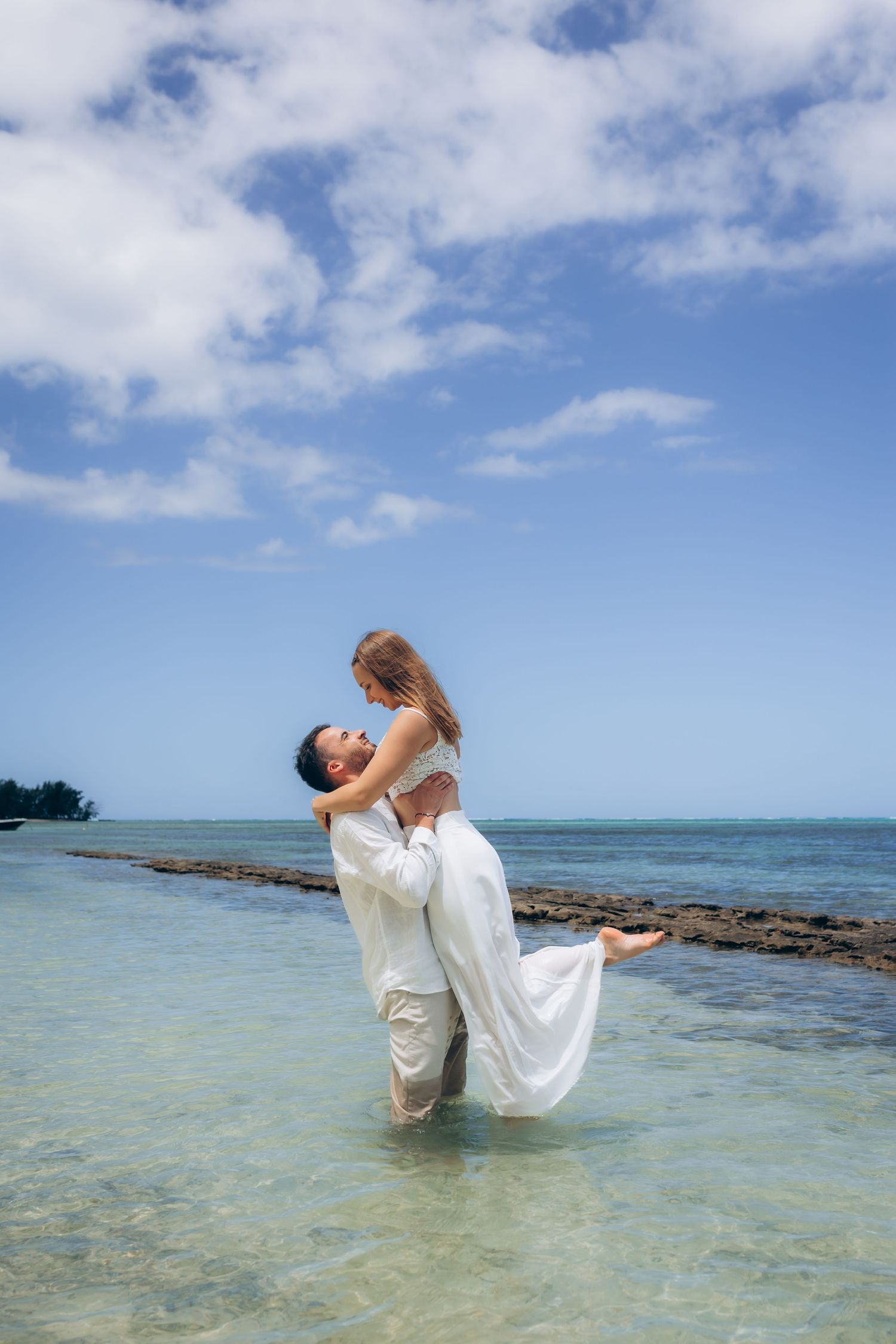 Le marié soulevant la mariée en robe blanche tout en se tenant dans l'eau peu profonde de l'océan tropical turquoise.