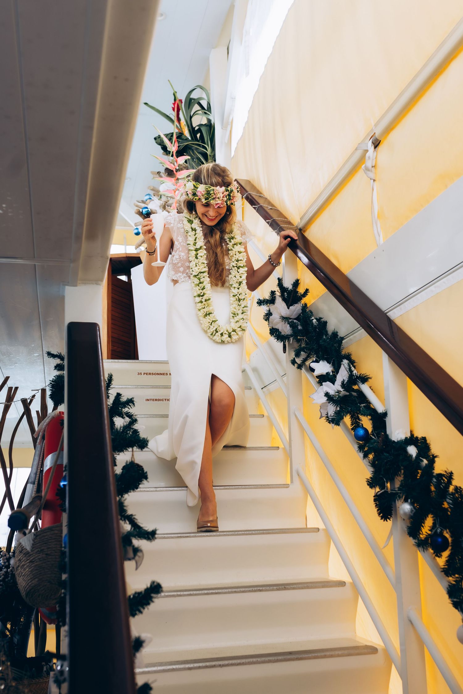 La mariée en robe blanche et lei à fleurs descend un escalier décoré dans un lieu de mariage tropical.
