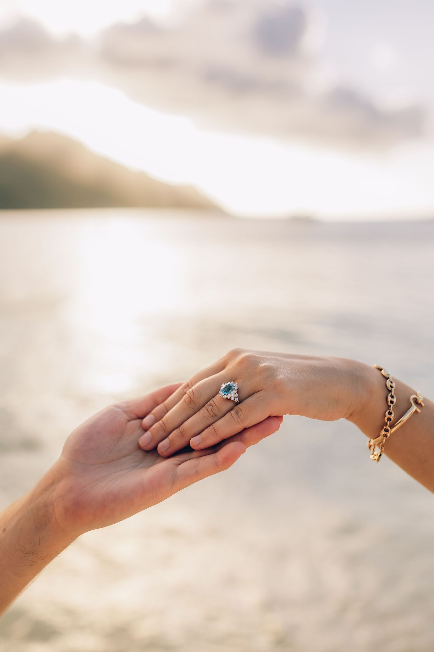 Deux mains se touchant sur une plage au coucher du soleil, l'une portant une bague de fiançailles halo saphir bleu et un bracelet en or.