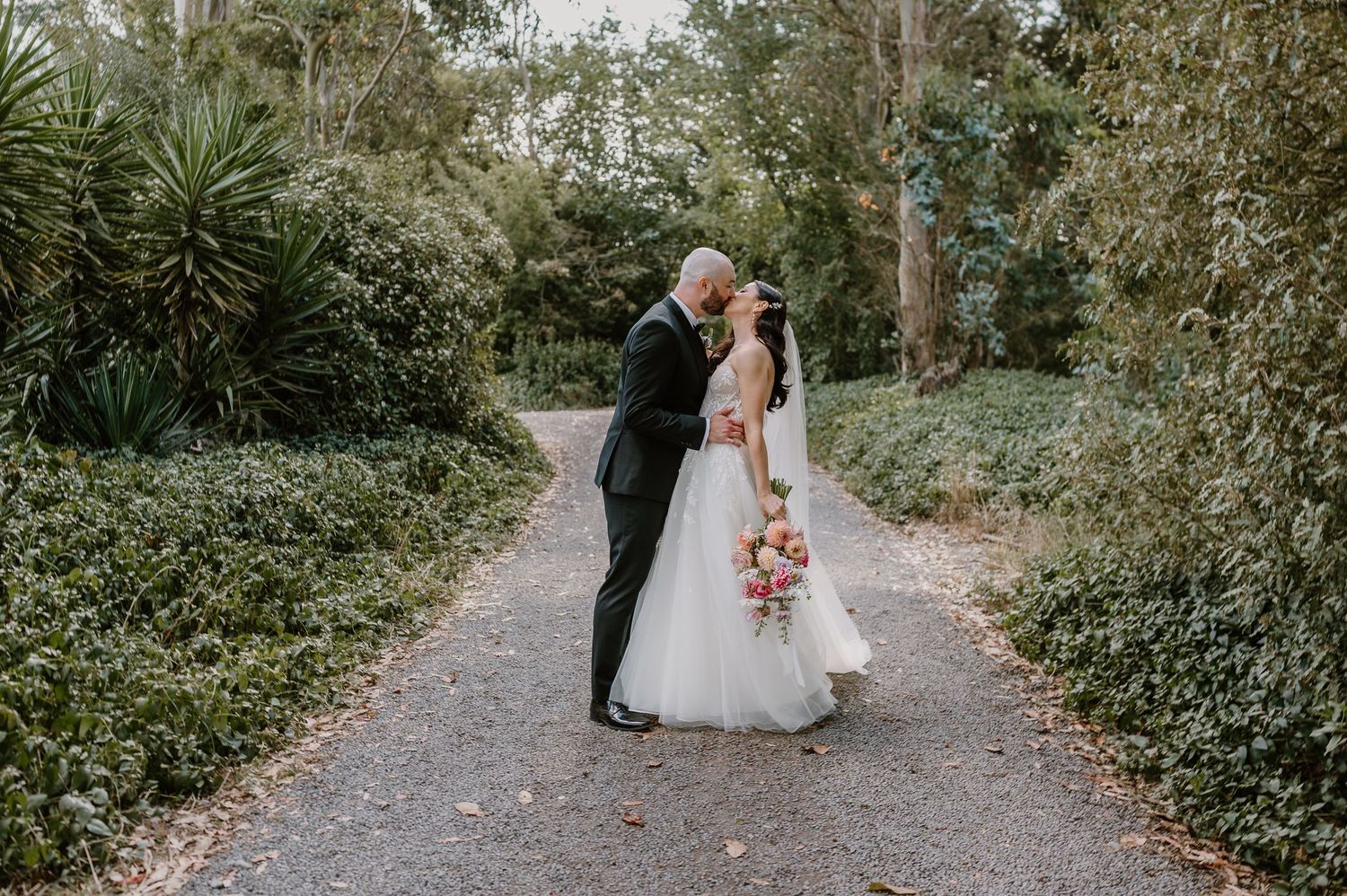 Two people share a romantic moment on a tree-lined garden path during a wedding ceremony.Trents Vineyard, Christchurch photographer
