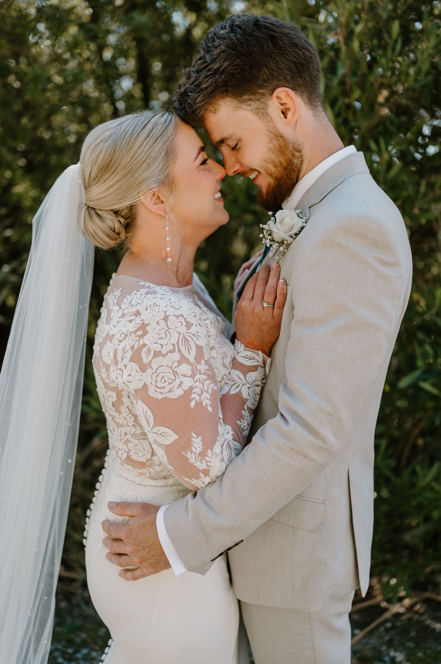 Newlywed couple shares intimate moment outdoors, bride wearing lace sleeve gown with veil and groom in light gray suit.