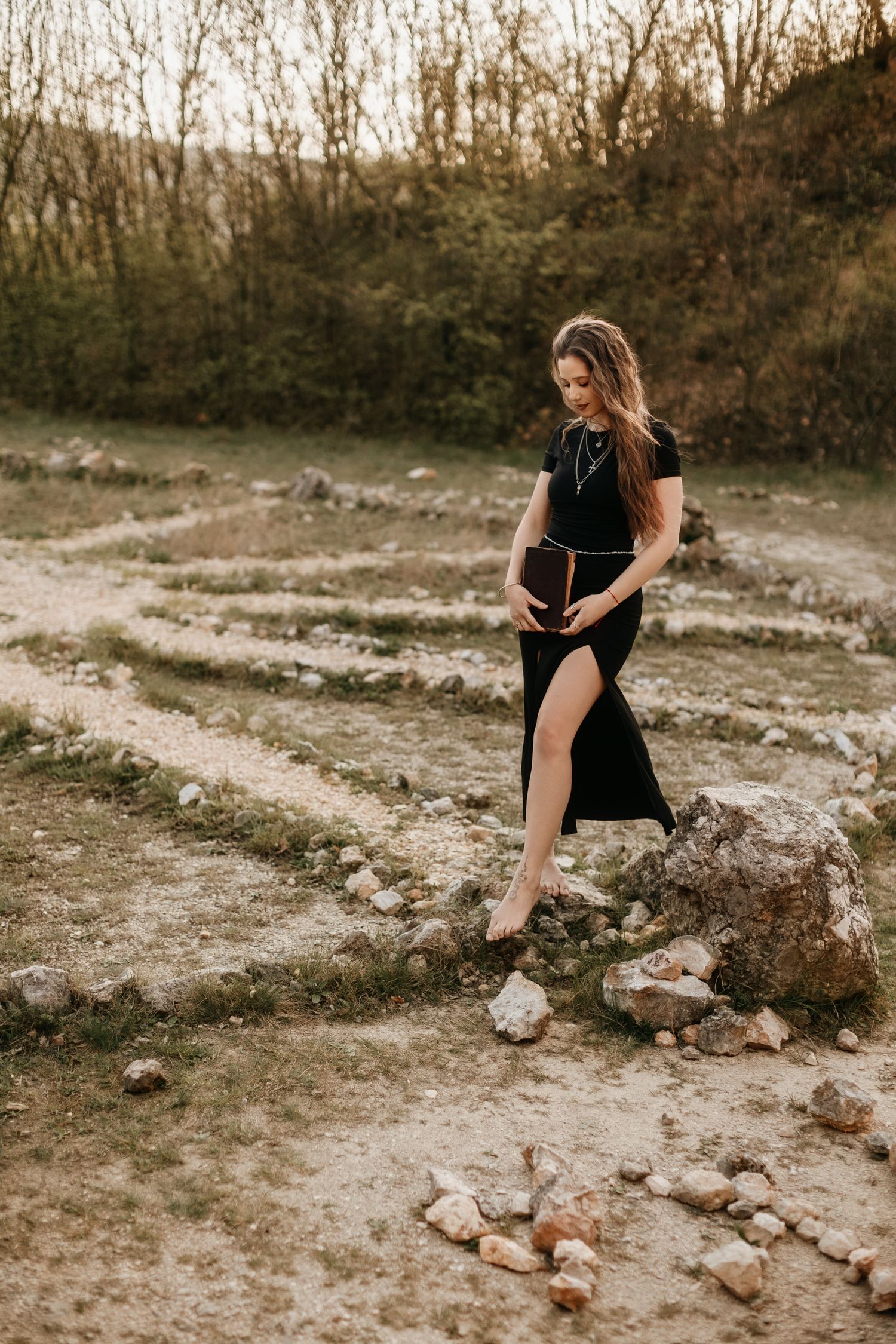 Fekete, lenge ruhát viselő nő áll egy kisebb sziklán, természetes kültéri háttér előtt.