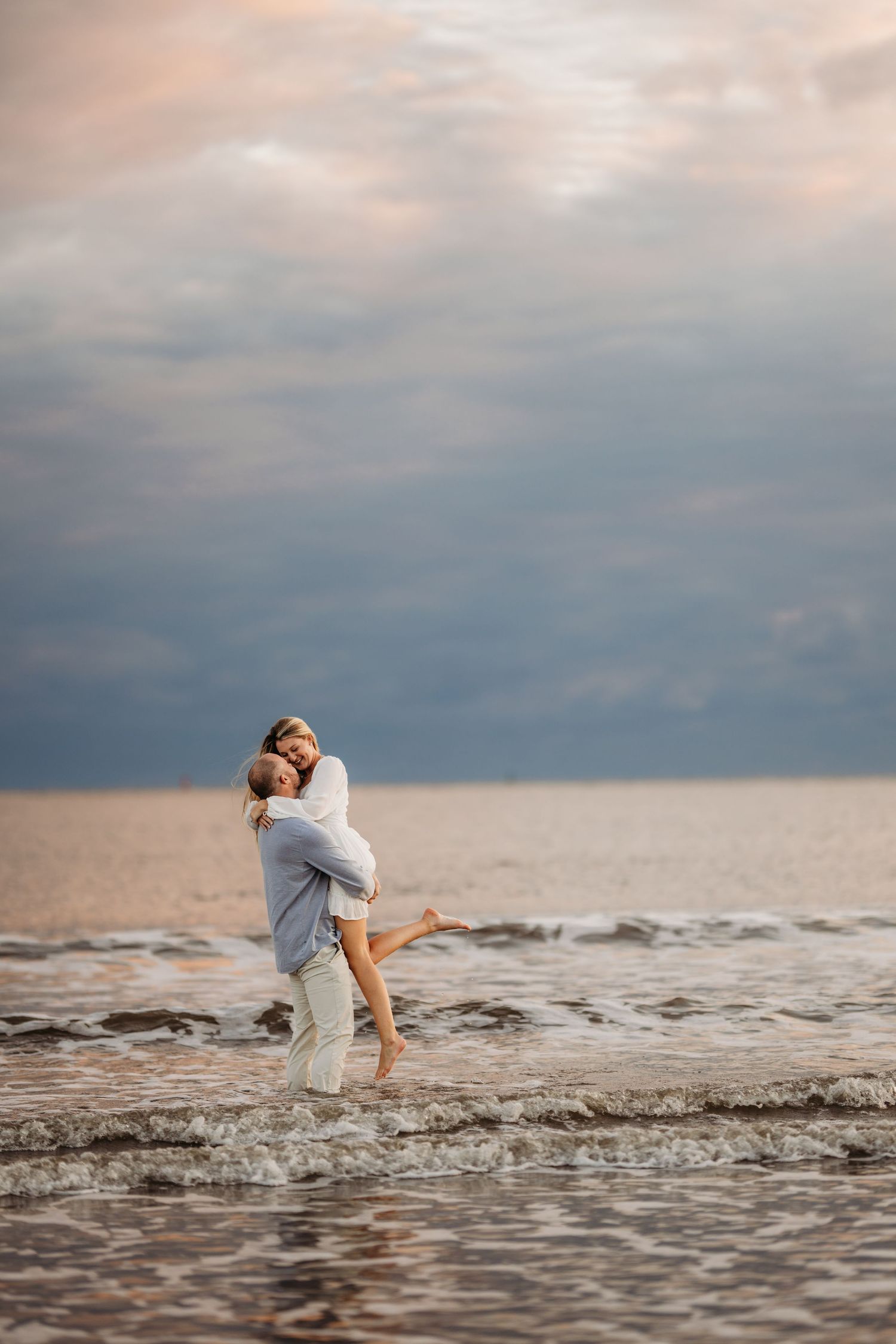 Joyful couple embracing in ocean waves during sunset with dramatic cloudy sky. Charleston, South Carolina.