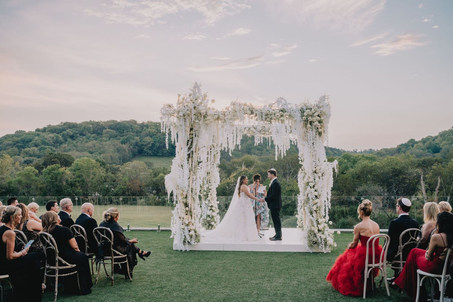 An outdoor wedding ceremony under a floral arch with guests seated on white chairs. Nashville, Tennessee.