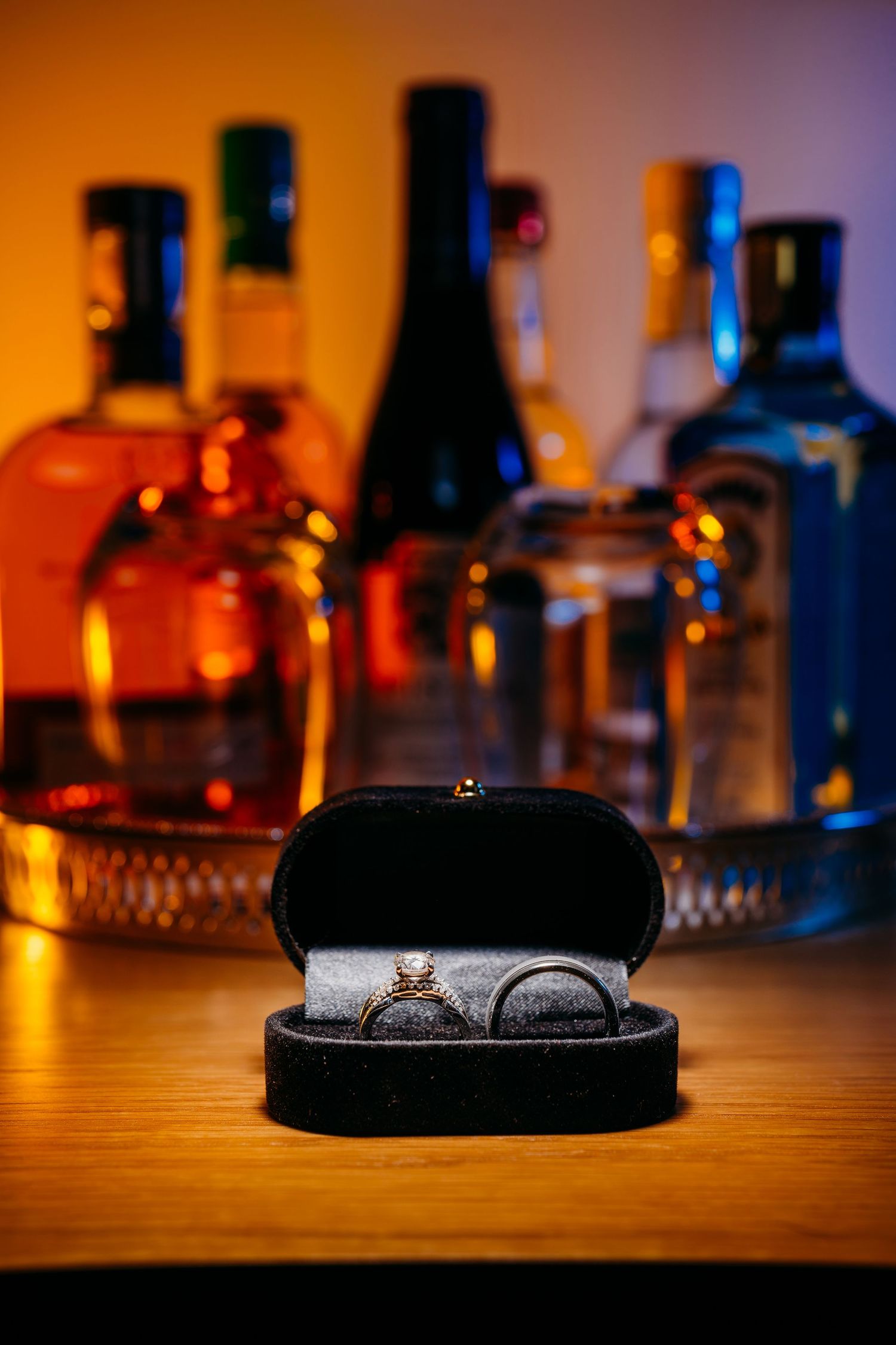 Wedding rings displayed in a black box with liquor bottles creating a warm ambient backdrop. Savannah, Georgia.