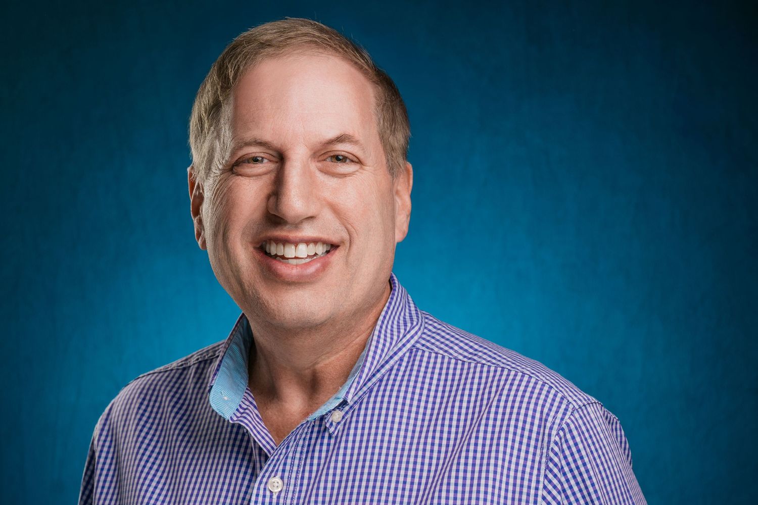 Professional headshot of person in blue checkered shirt smiling against blue backdrop. Nashville, Tennessee.