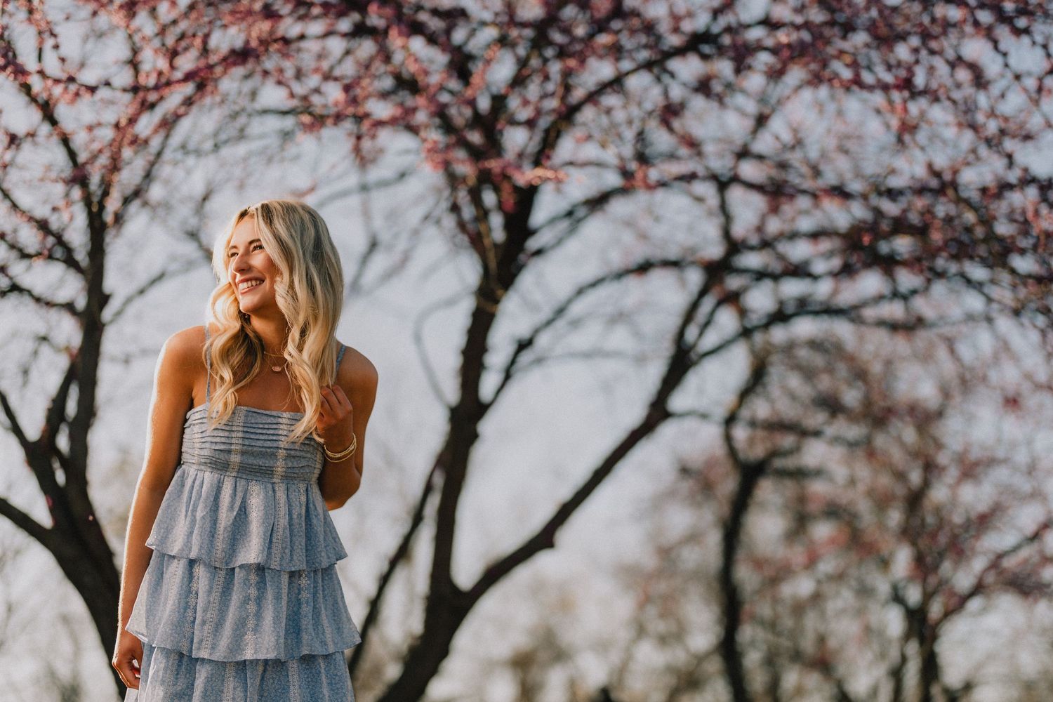 A dreamy portrait in a tiered blue dress stands against blooming cherry trees in soft lighting. Murfreesboro, Tennessee.