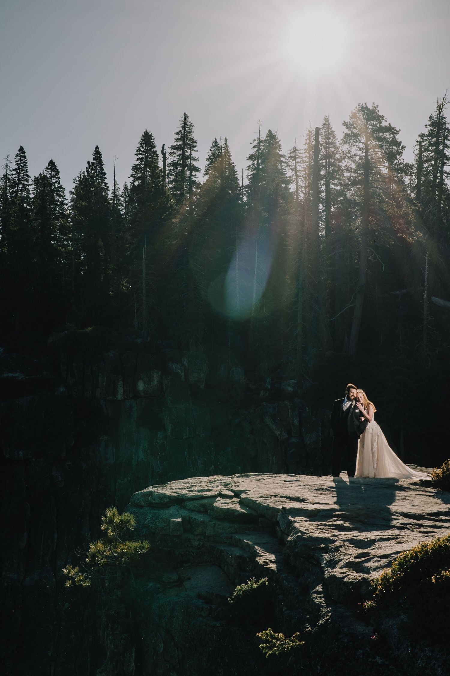 Wedding couple standing on rocky cliff edge with sunlit pine forest backdrop. Yosemite national Park, California.