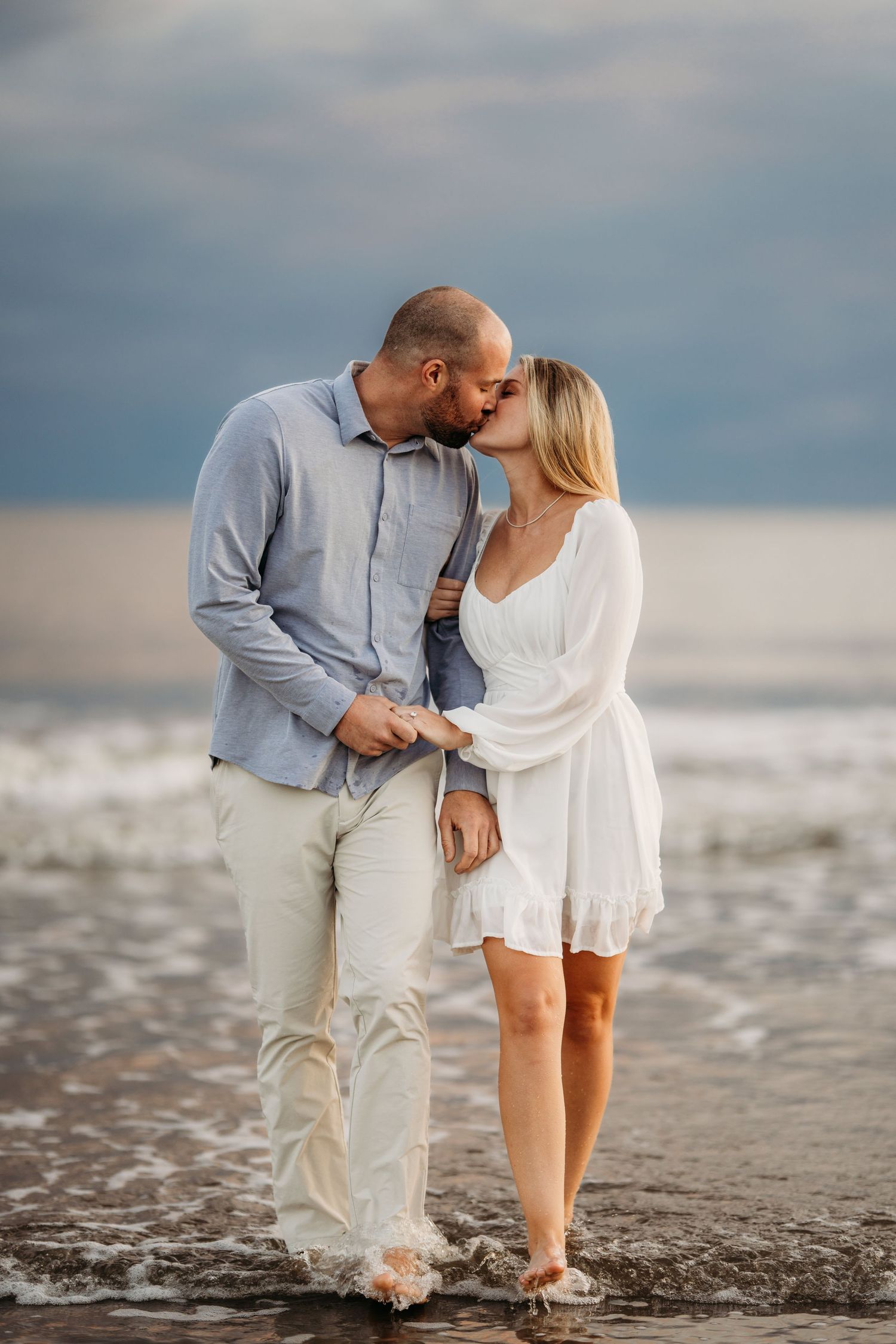 Romantic couple kissing while standing in shallow ocean waves during moody sunset. Charleston, South Carolina Carolina.