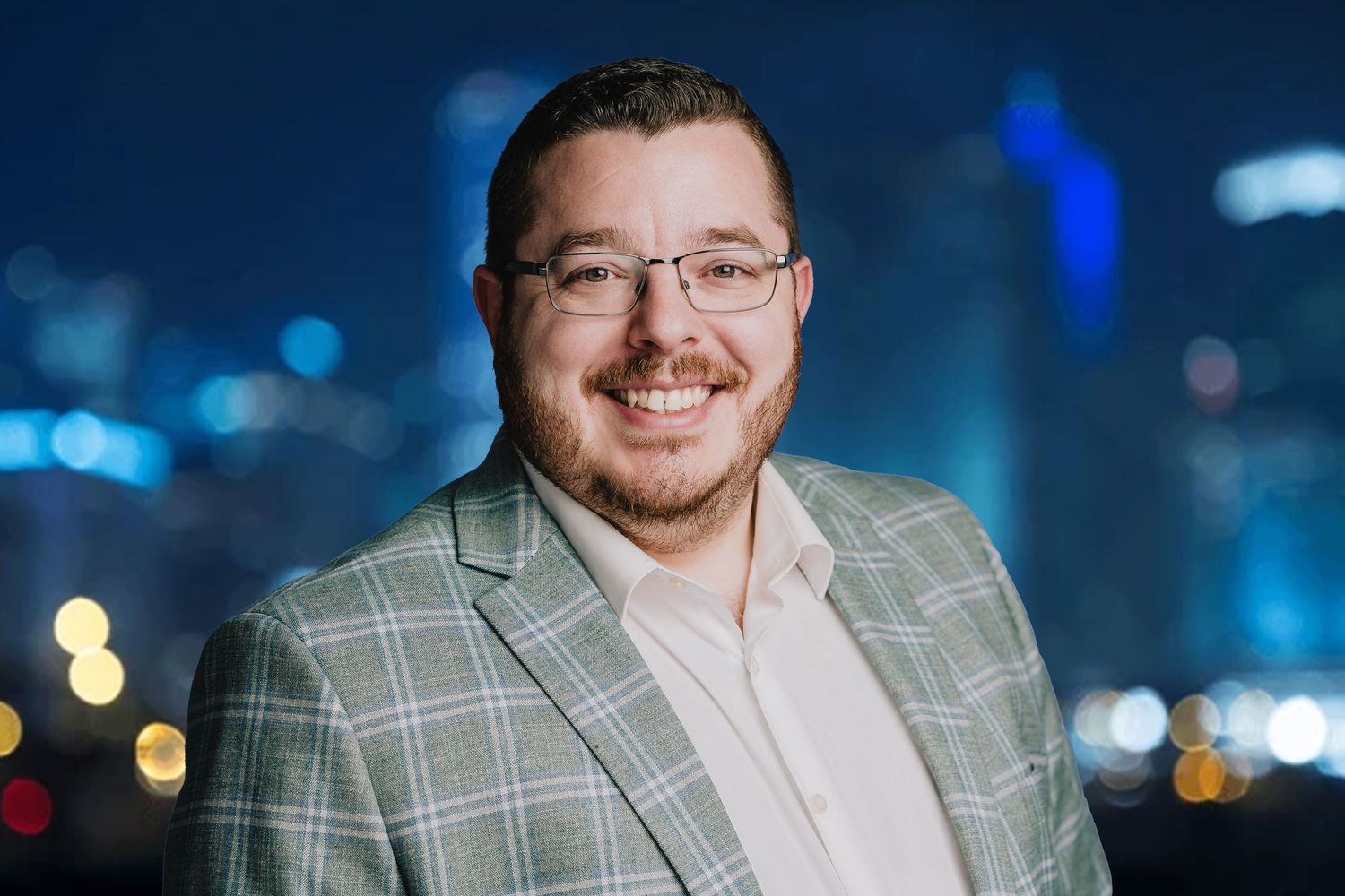 Person in plaid blazer and glasses smiling with city lights in background. Nashville, Tennessee.