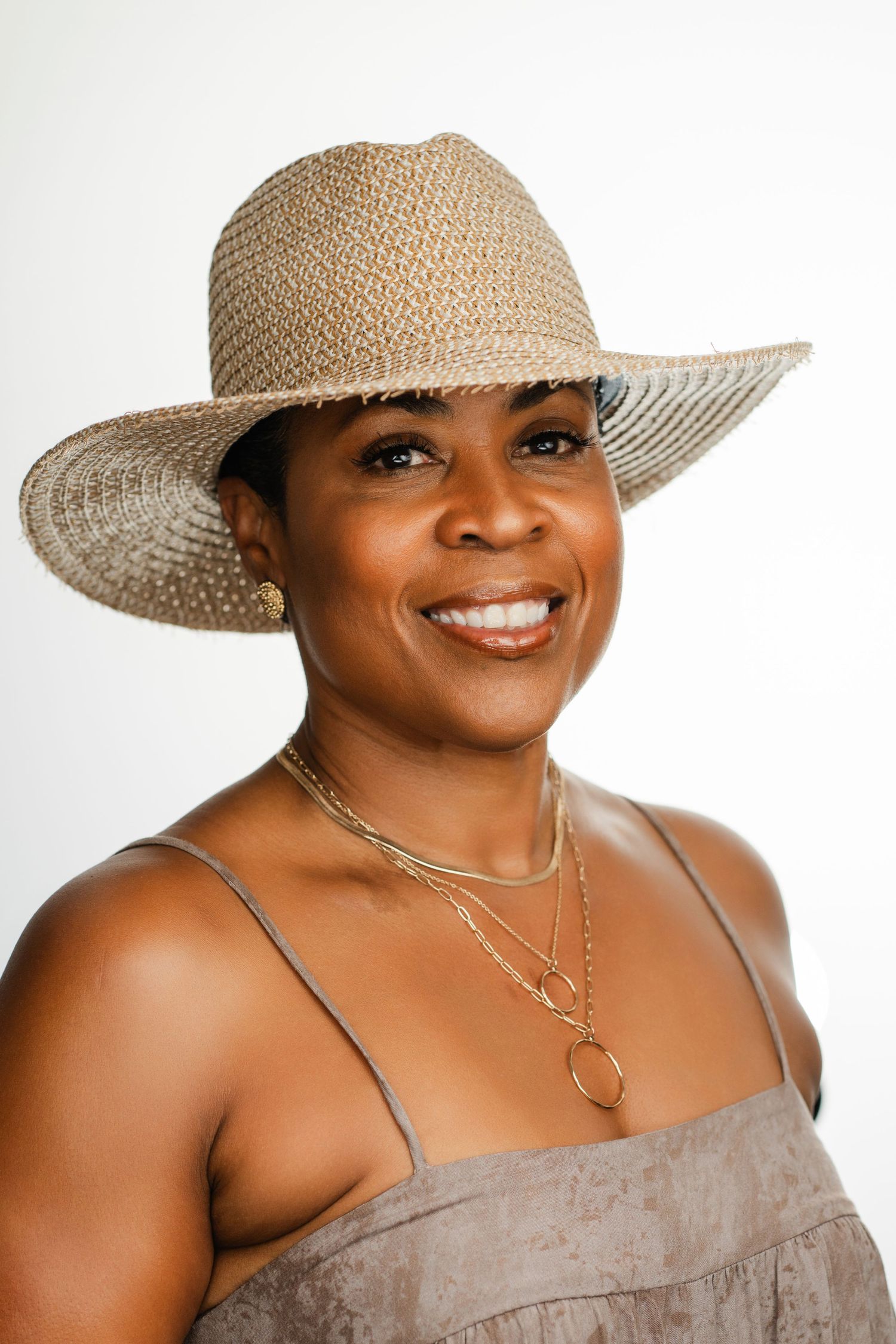 A warm smile radiates from beneath a woven sun hat, complemented by layered necklaces and a brown dress. Nashville, Tennessee.