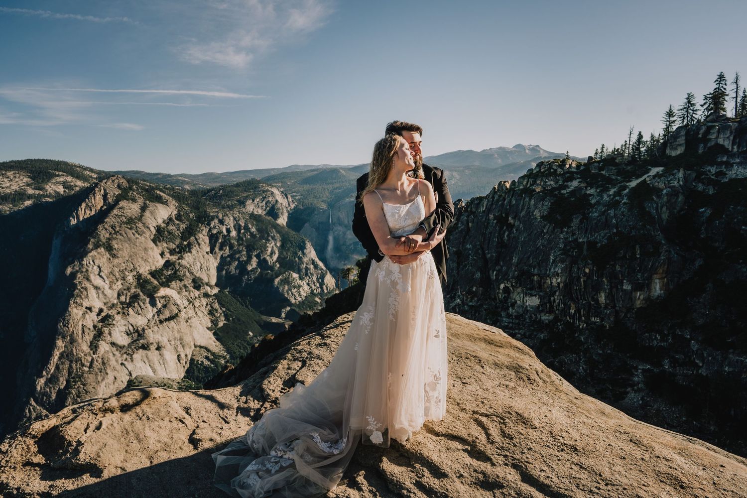 Bride in flowing dress standing on mountain peak overlooking dramatic valley vista. Yosemite national Park, California.