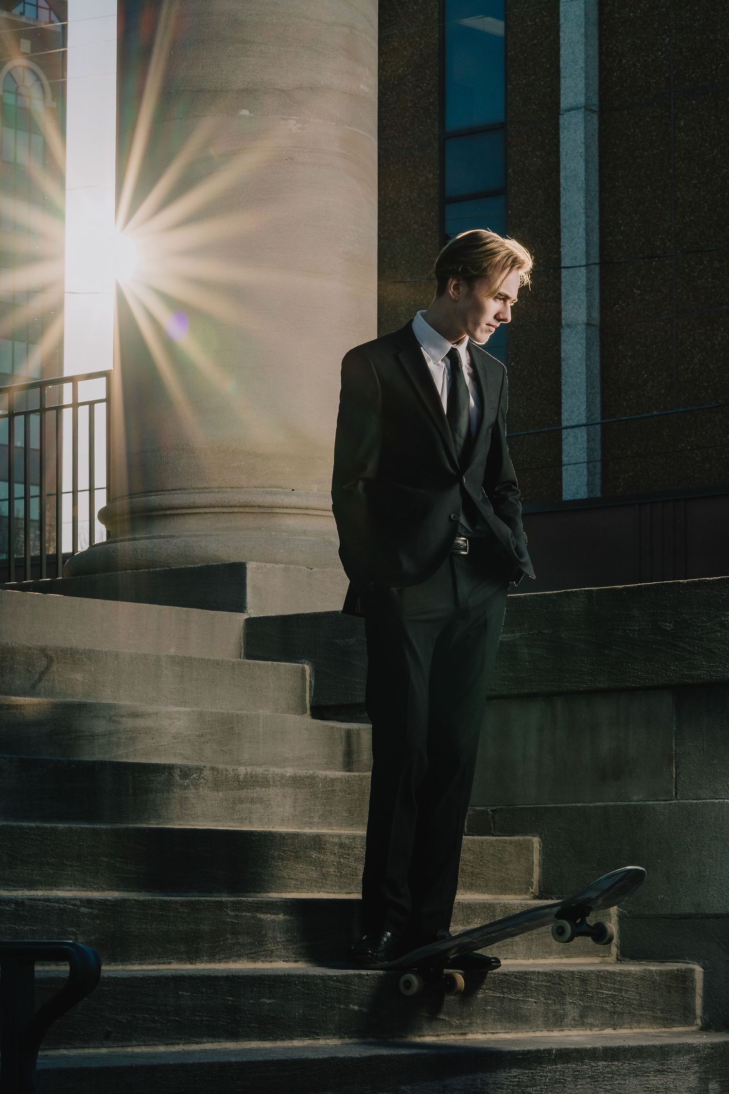 Professional in black suit poses with skateboard on stone steps as sunlight creates dramatic starburst effect. Nashville, Tennessee.
