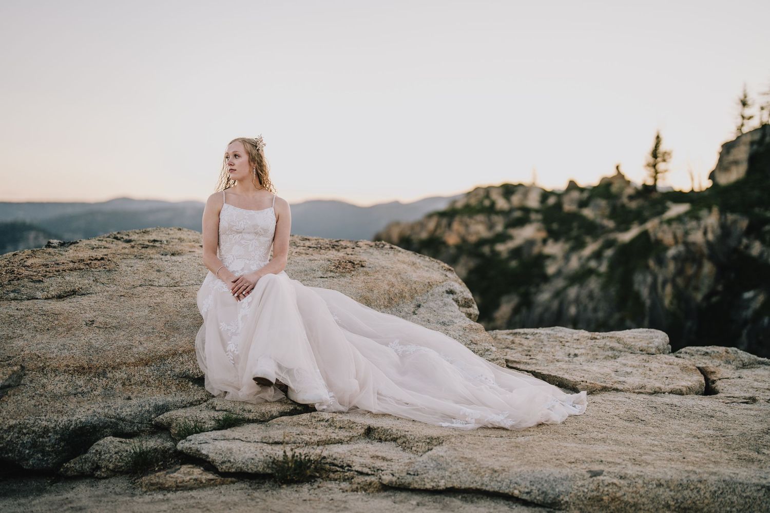 Bride in white dress sitting on rocky cliff overlooking mountainous landscape at sunset. Yosemite national Park