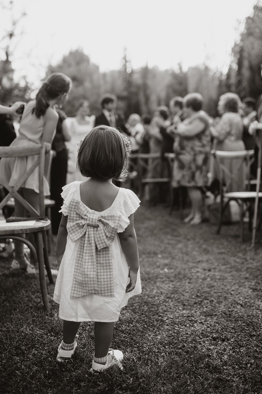 A child in a dress with gingham bow watches wedding festivities from behind.
