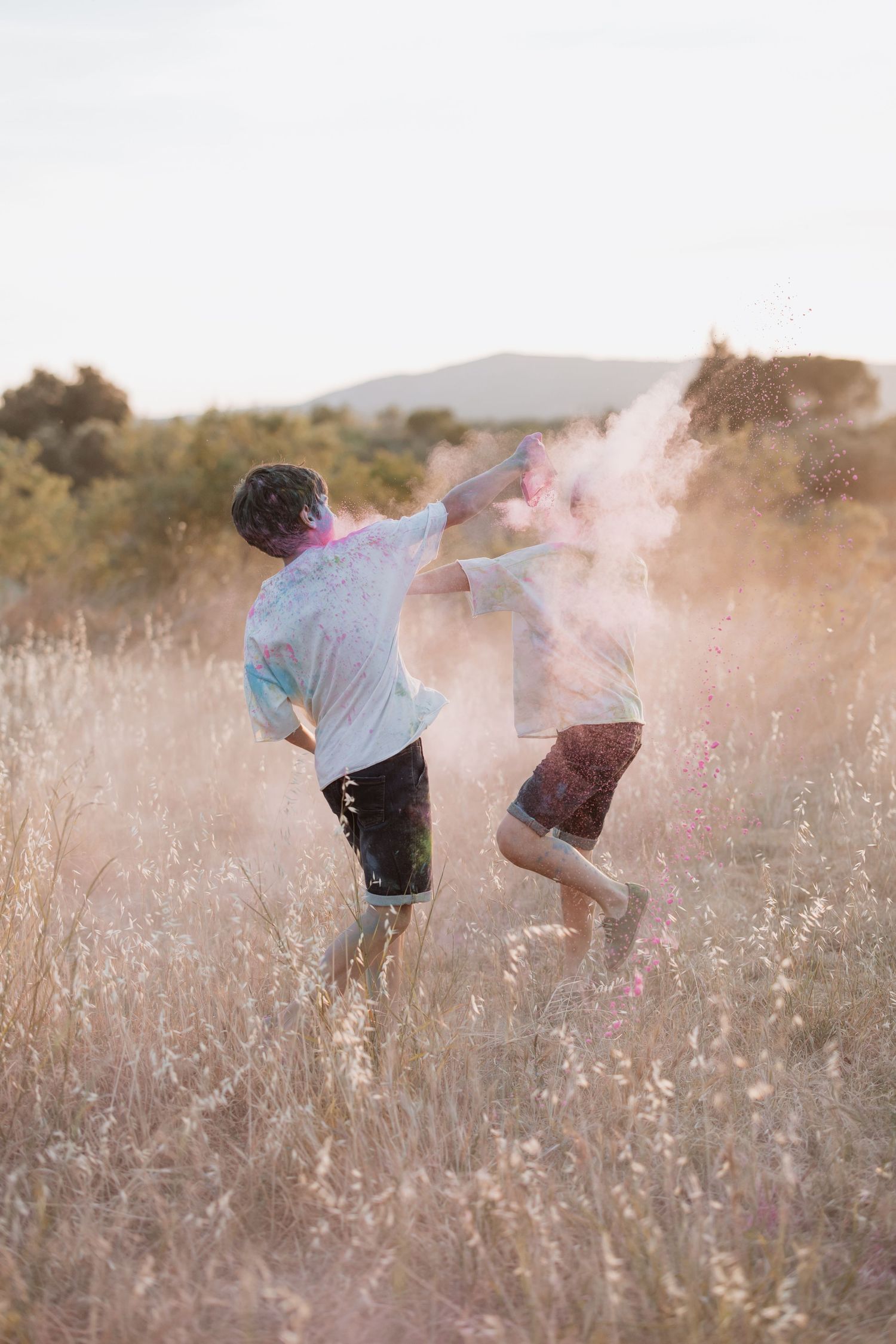 Two people playing with colorful powder in a golden meadow at sunset creating dreamy atmosphere in the summer field.