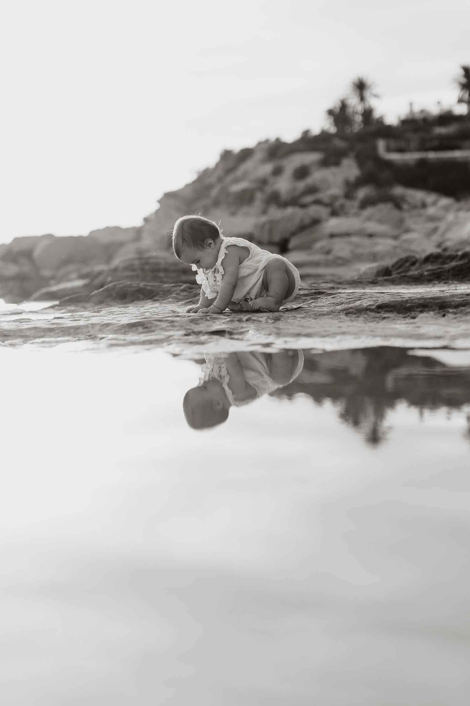 Black and white image of baby exploring water reflection on rocky beach.