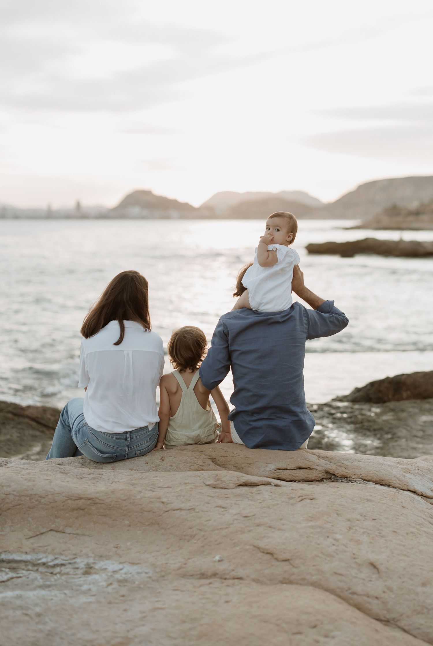 Family sitting together on coastal rocks overlooking the ocean and mountains at dusk.
